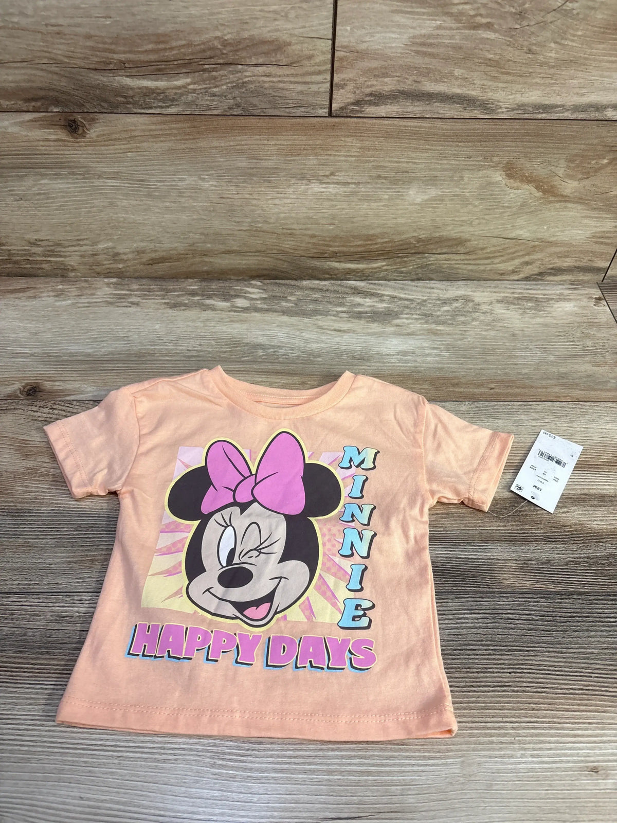 Flat lay of peach Minnie Mouse toddler shirt with winking Minnie face, pink bow, shows MINNIE and HAPPY DAYS on wood background
