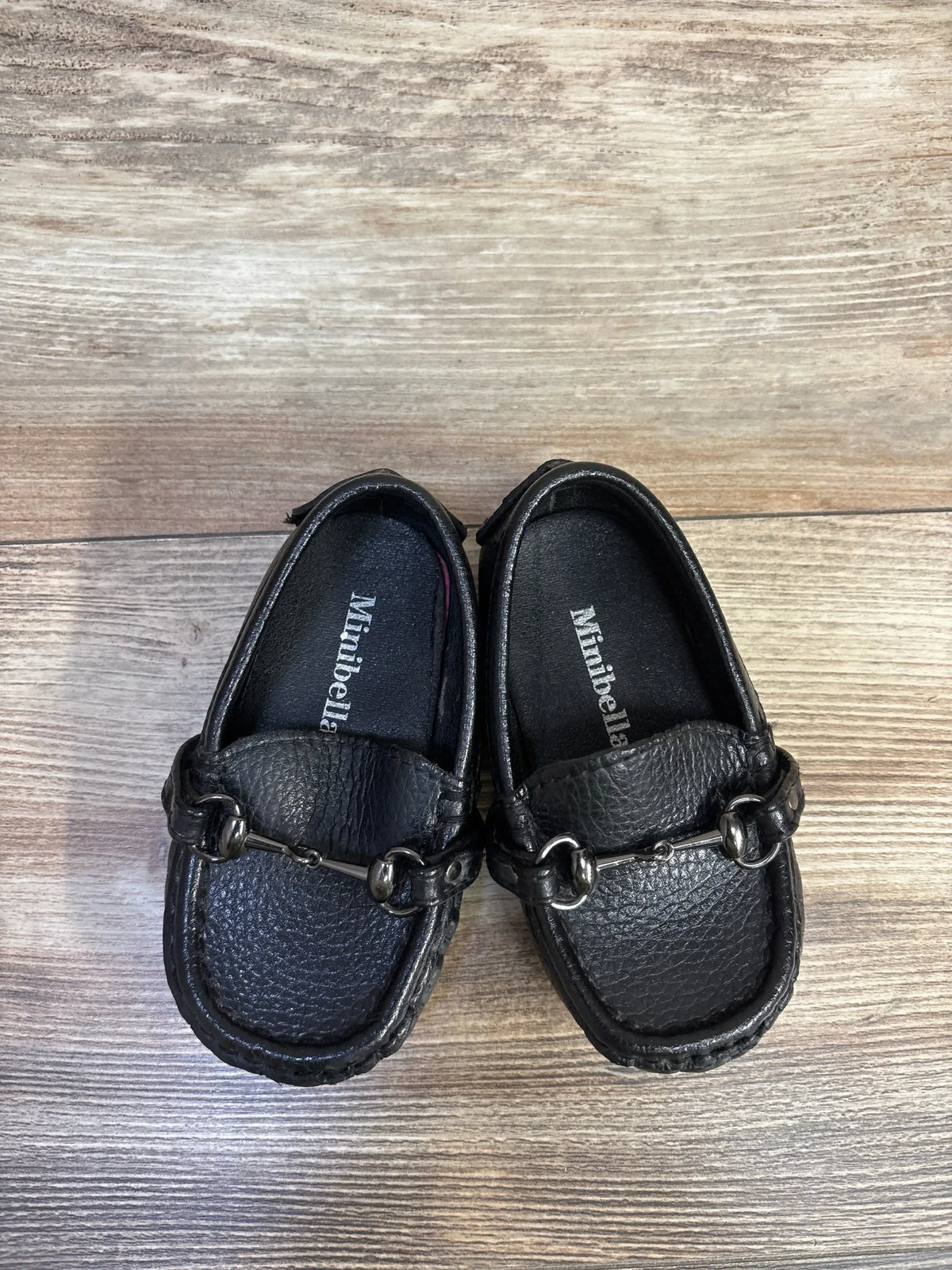 Top-down view of Minibella black leather slip-on toddler boat shoes showing metal bit and pebbled finish on wood floor