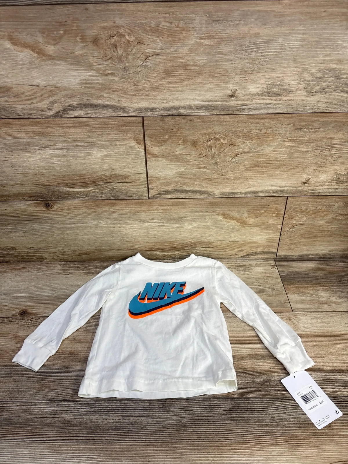 Back view of white toddler Nike long-sleeve shirt size 2T showcasing large blue and orange NIKE logo laid flat on wood floor