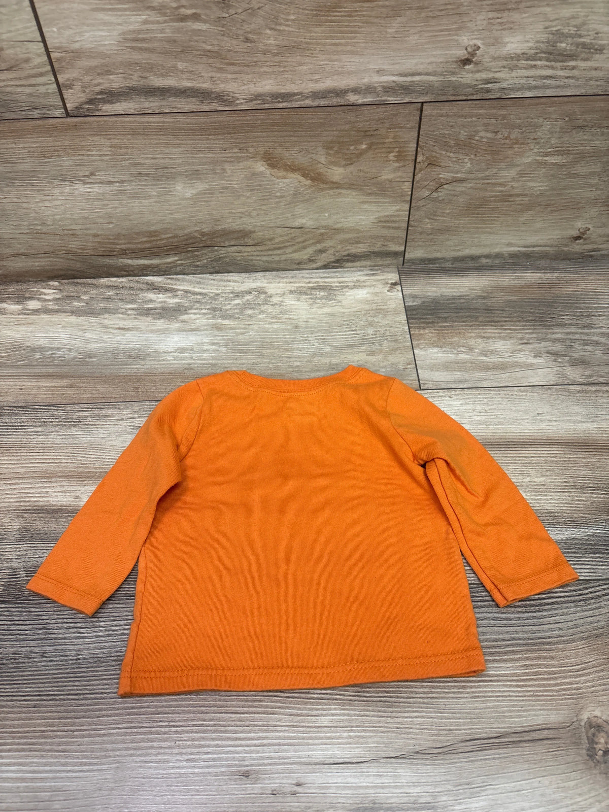 Top view of an orange long-sleeve baby shirt showcasing its plain front on a wood-look floor