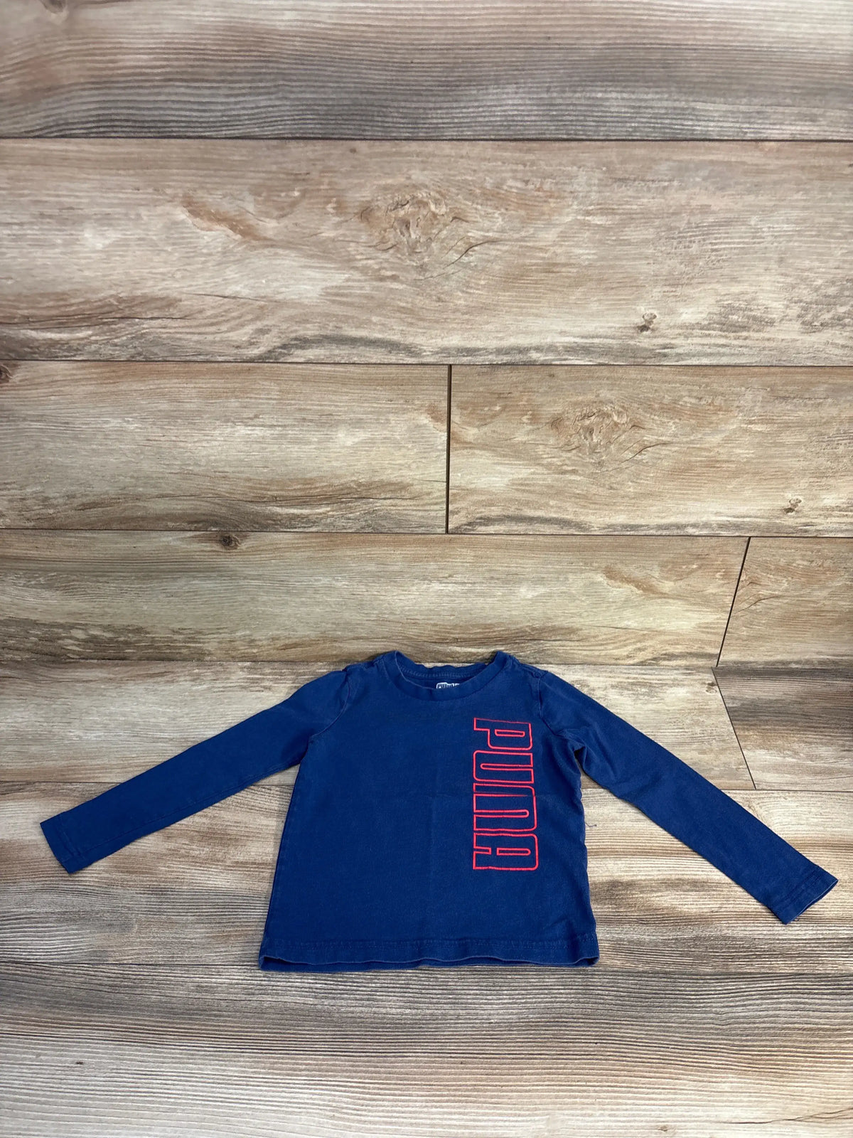 Front view of a navy Puma 3T toddler shirt showcasing long sleeves and red Puma lettering on a wood plank background