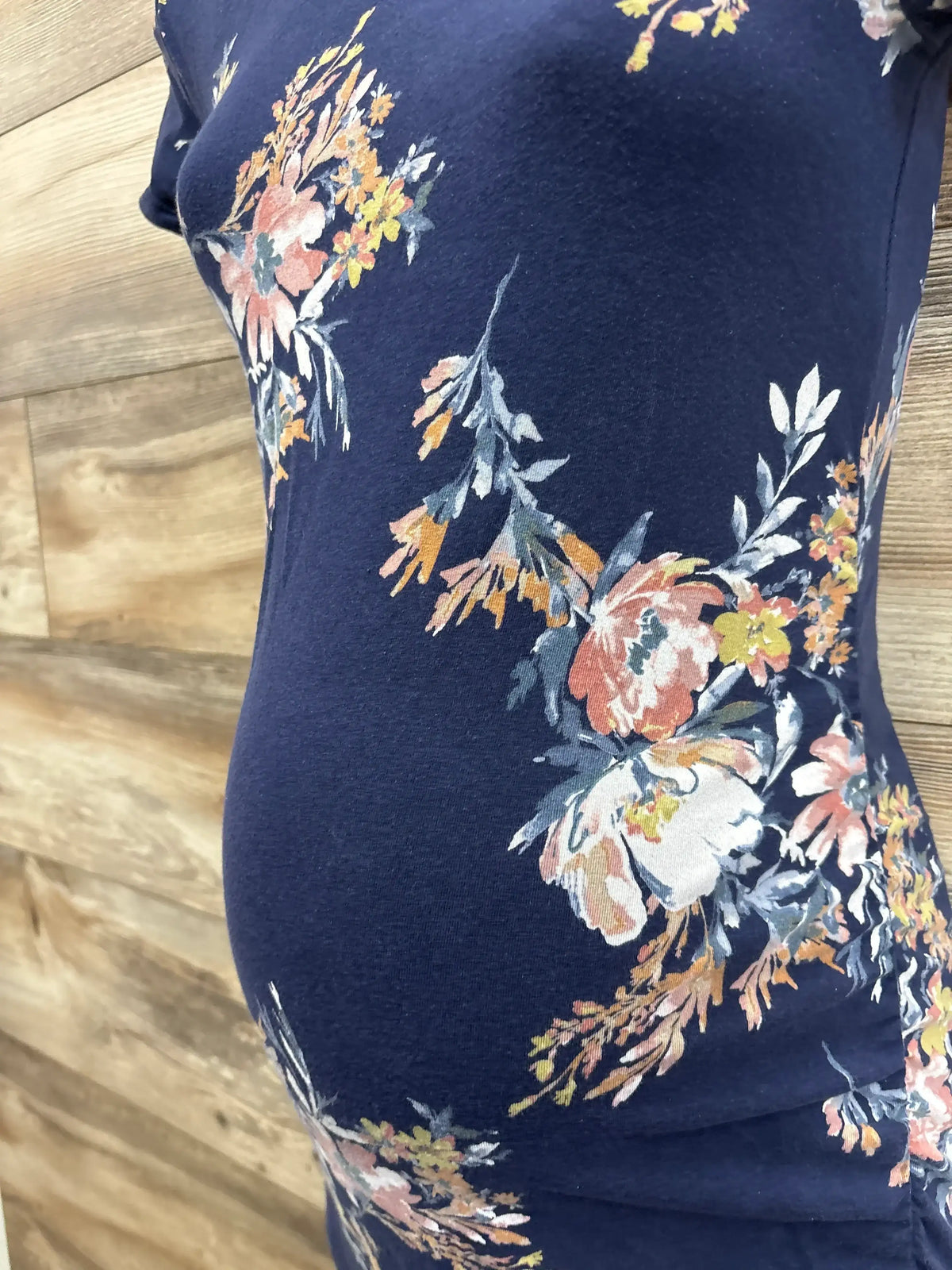 Side close up of navy Isabel maternity bodycon dress on mannequin showing floral print and ruched belly with wood background