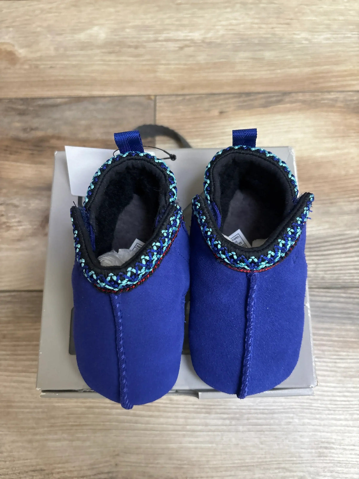Blue infant baby shoes with decorative trim, displayed on a box, wooden floor background