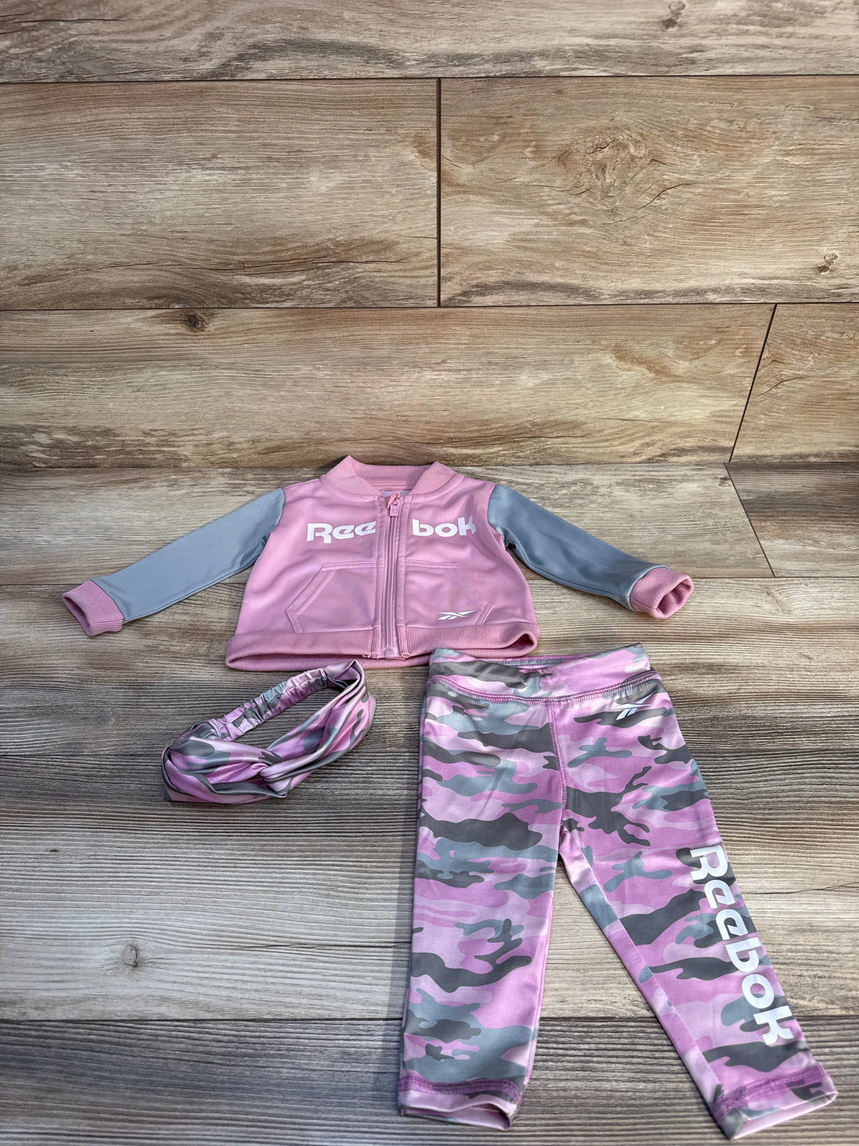 Pink and gray Reebok toddler athletic outfit with jacket, leggings, and matching headband on wood floor
