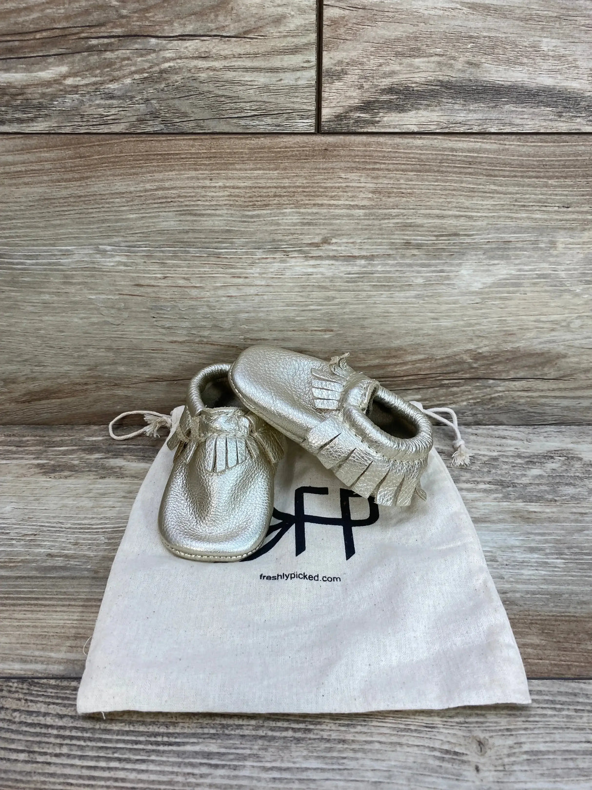 Angled front view of platinum Freshly Picked baby moccasin size 2 showcasing soft leather and fringe on canvas bag and wood backdrop