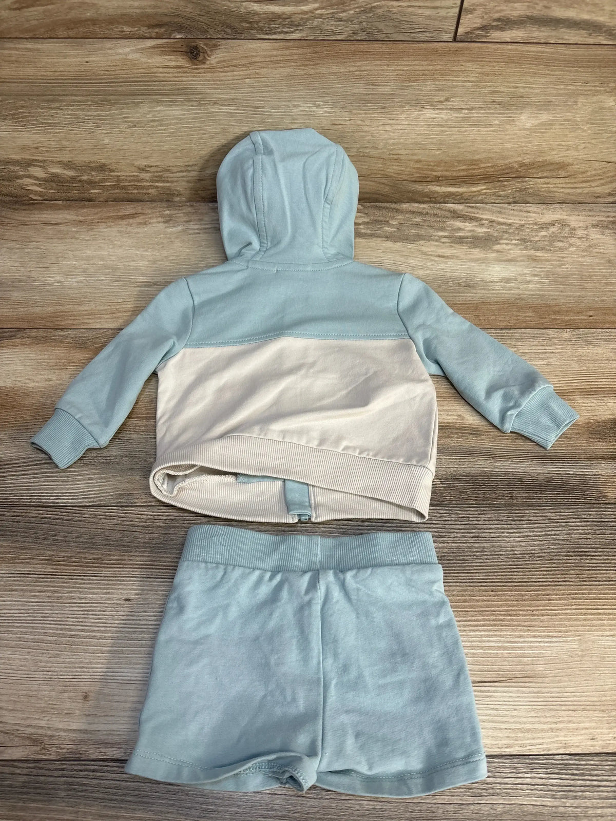 Back view of Max Studio baby 2-piece zip-up hoodie and shorts showing light blue hood and cream color-block panel on wood floor