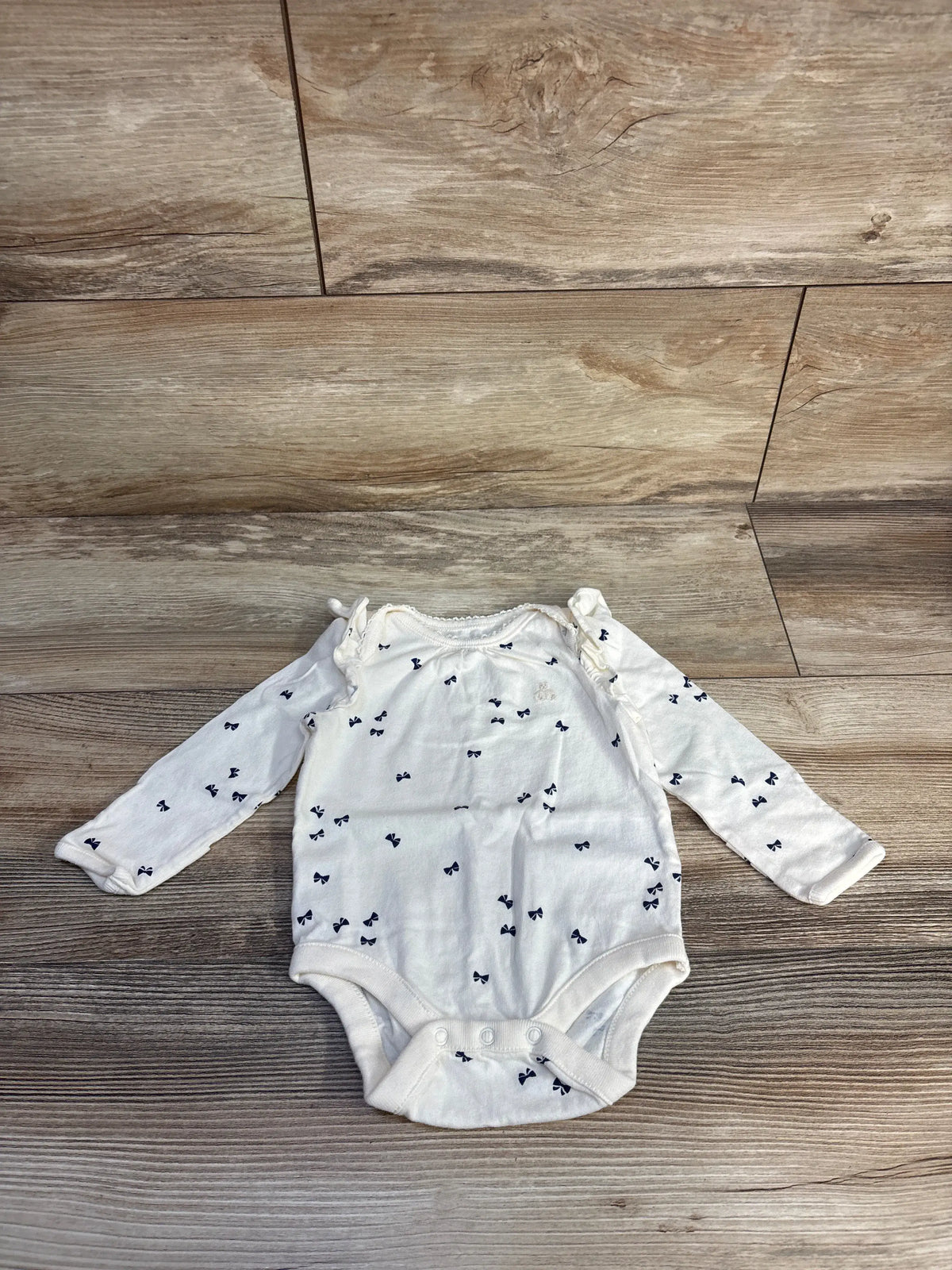Front view of Baby Gap cream long sleeve bodysuit showcasing navy bow print, fold-over hand cuffs, and snap closures on wood floor