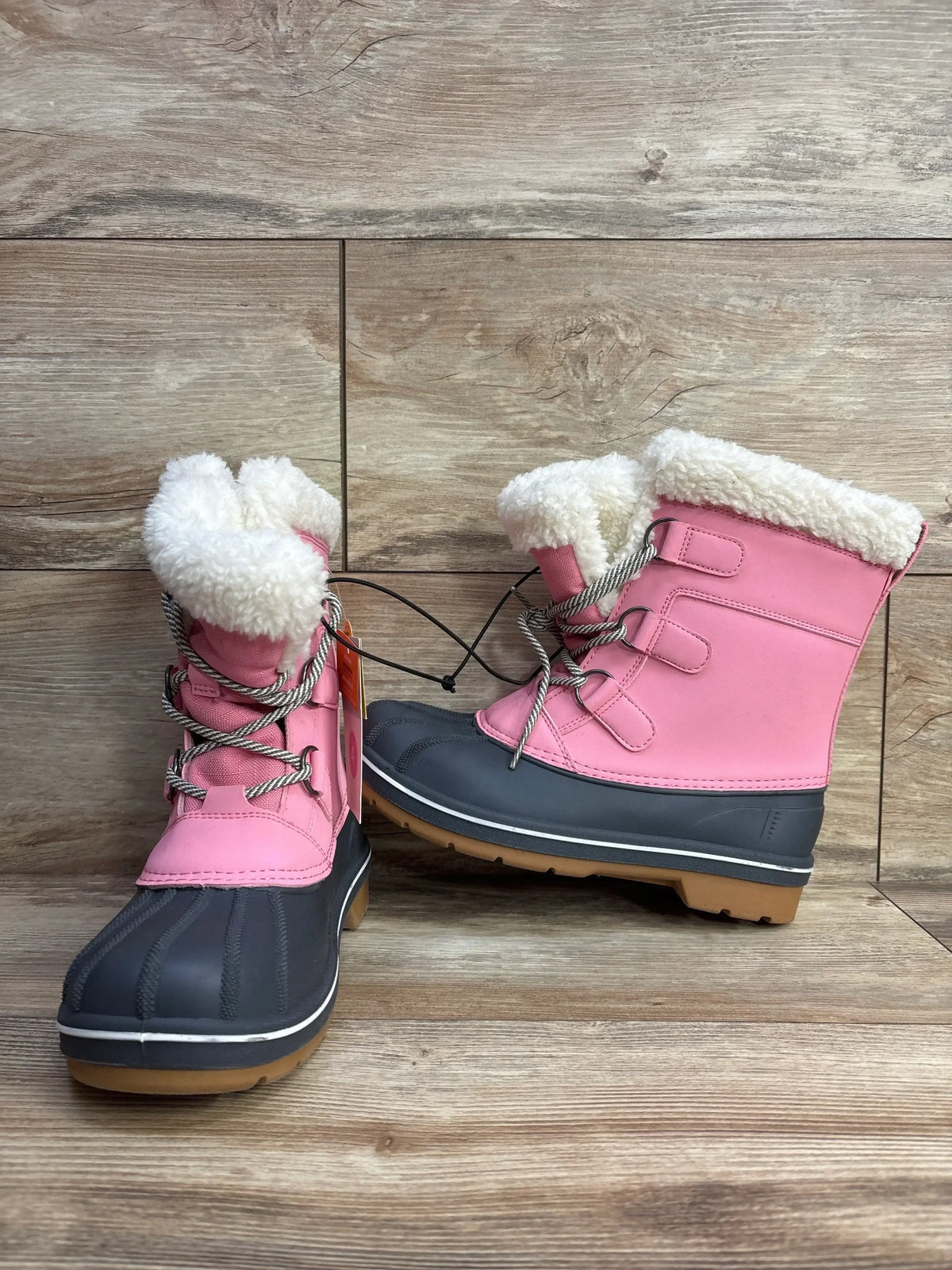 Three quarter view of pink kids winter snow boots with cream faux fur cuffs lace up fronts and black rubber soles on wood plank floor