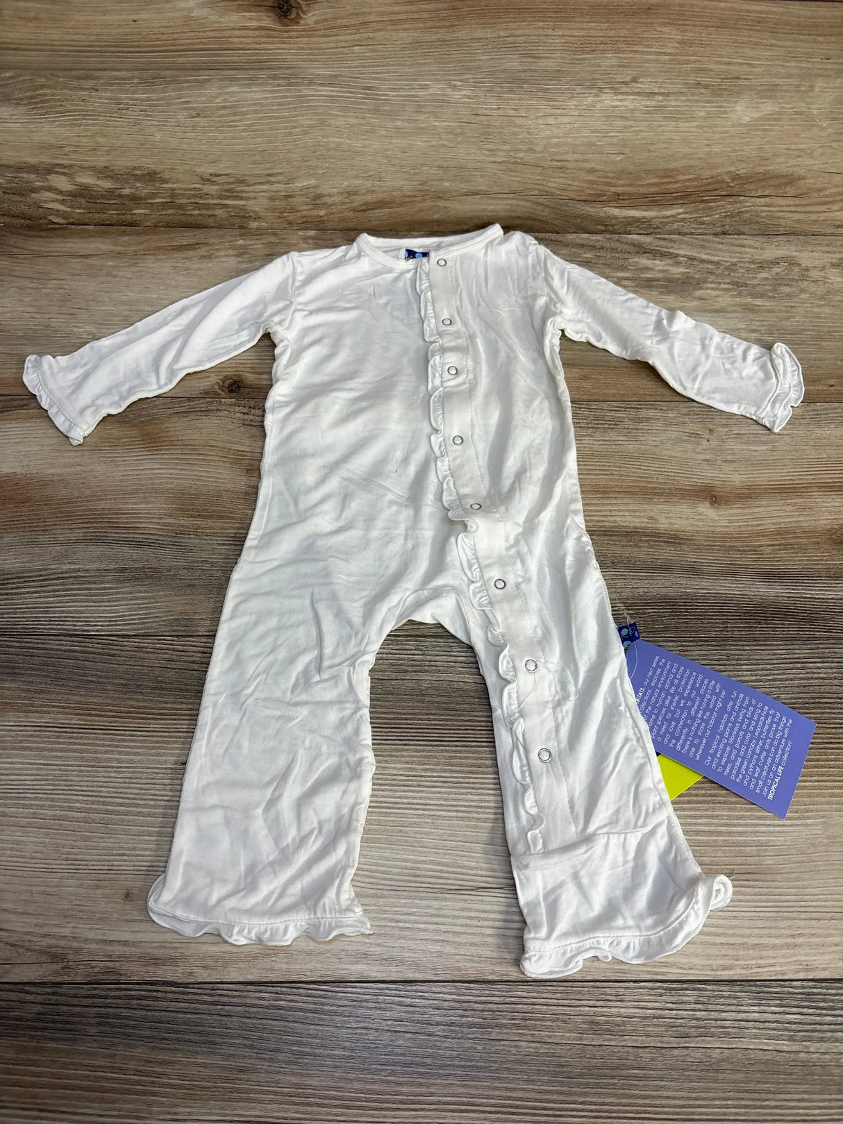 Front view of white KicKee Pants ruffle coverall size 6-9 months showing front snap closure and ruffle trim, laid flat on wood background