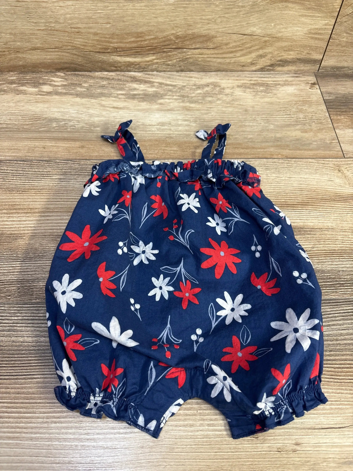 Flat lay of blue floral tank shortie romper size 3M with red and white flowers, knotted straps and elastic legs on wood