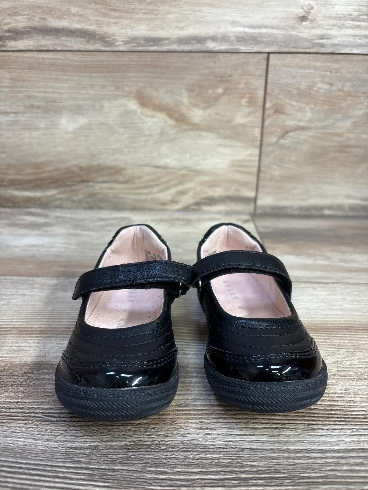 Front view of Kelly and Kate Mary Jane flats showing black patent toe, Velcro strap and rubber sole on wood floor