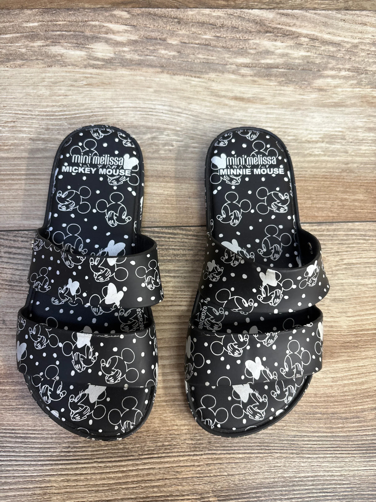 Top view of Mini Melissa x Disney Minnie Mouse black slides showing white Mickey and Minnie print on dual straps on wood floor