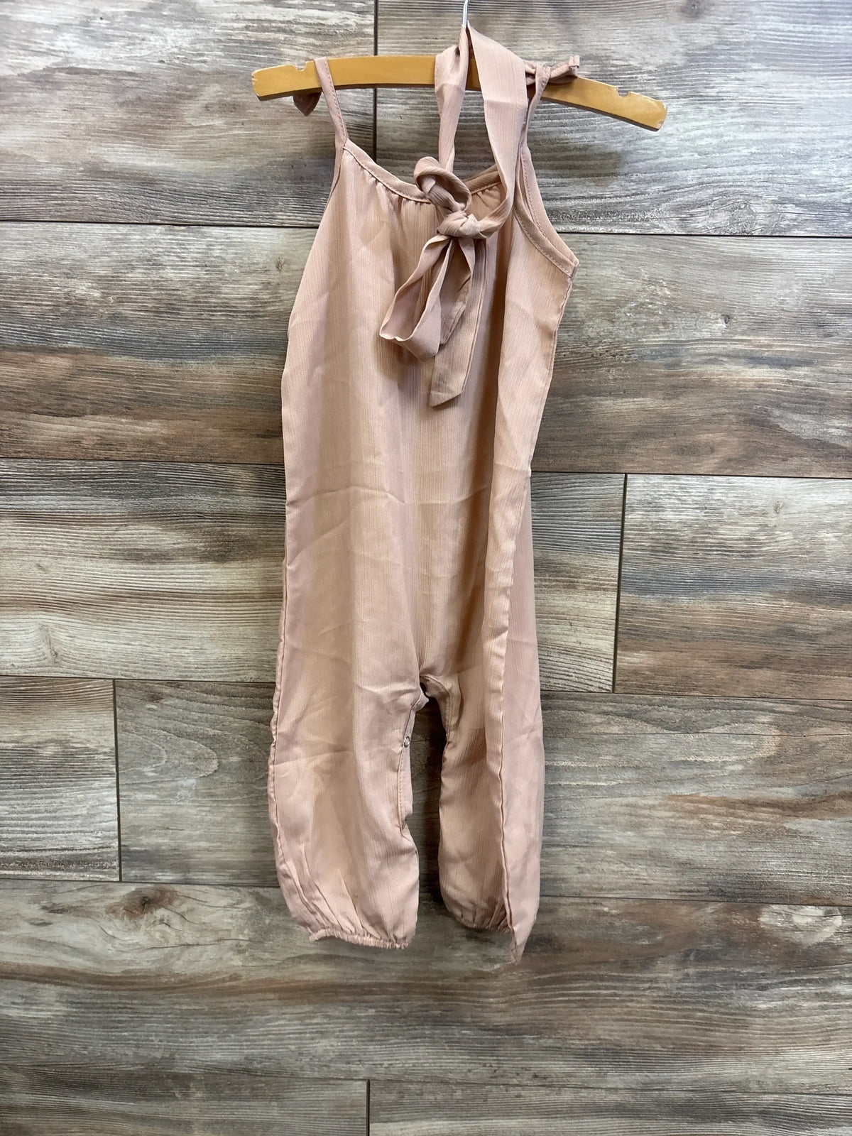 Front view of beige baby jumpsuit with halter tie and elastic ankle cuffs on wooden hanger against wood background