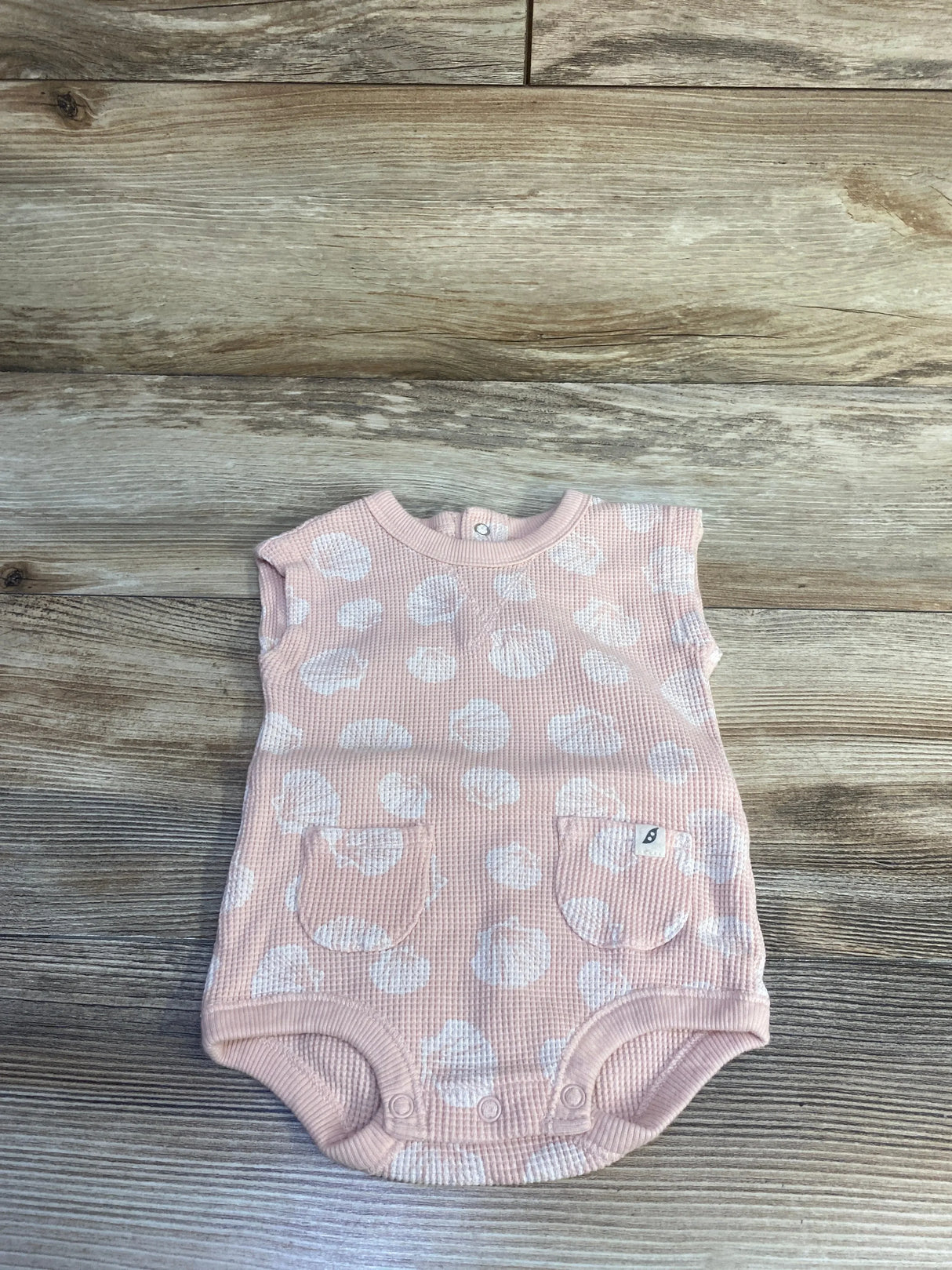 Top-down view of peach waffle-knit baby tank bodysuit (size 3–6m) with white polka dots, two front patch pockets, and crotch snaps on wood plank background