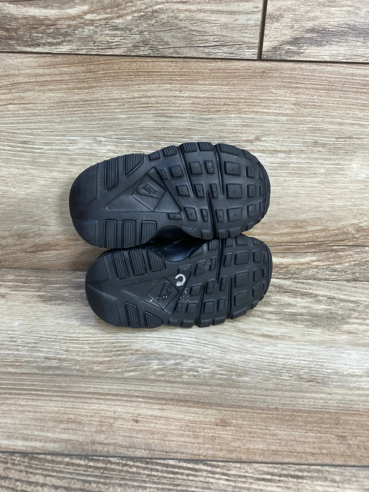 Bottom view of toddler Nike Air Huarache Triple Black sneakers showing black rubber soles and tread pattern on wood floor