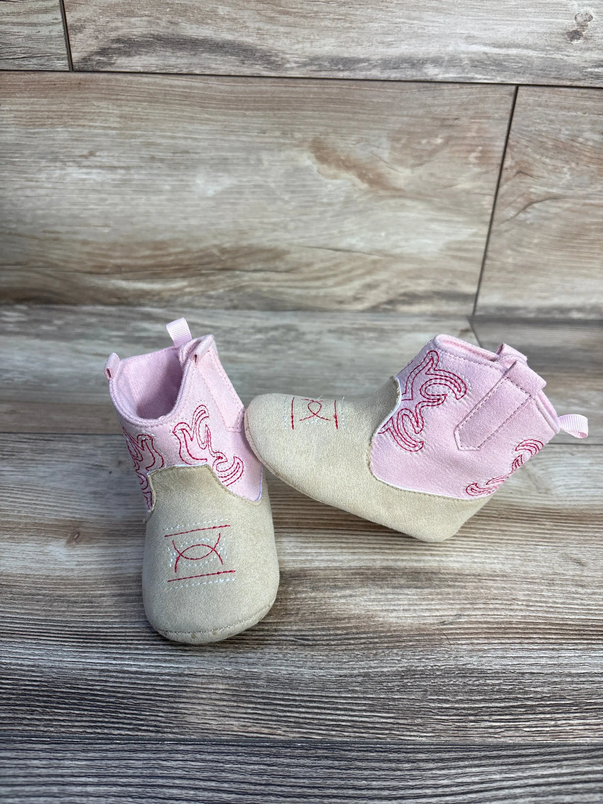 Front and side views of Stepping Stones cowgirl crib boots showcasing pink embroidered uppers and cream toes on a wood floor