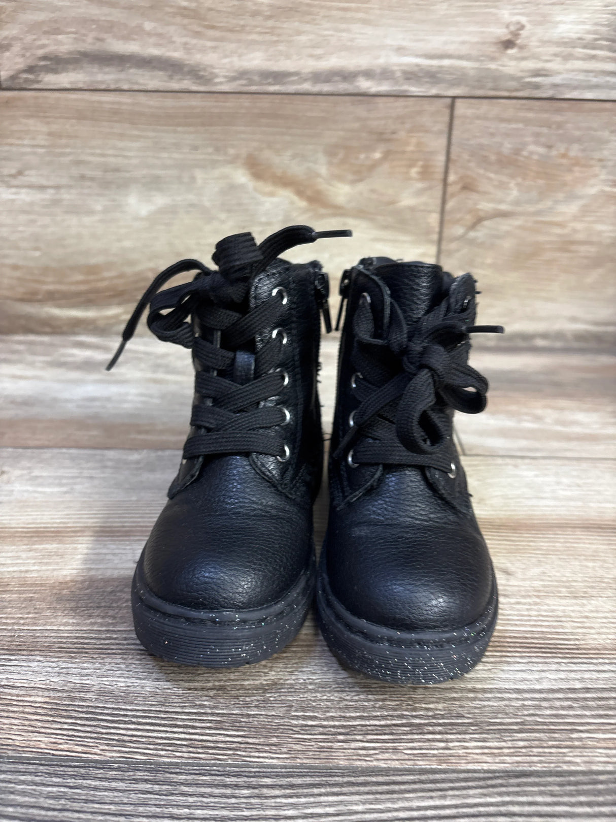 Front view of Sincerely Jules toddler black glitter lace-up boots showing glitter-speckled soles, side zippers, and textured leather on wood floor background