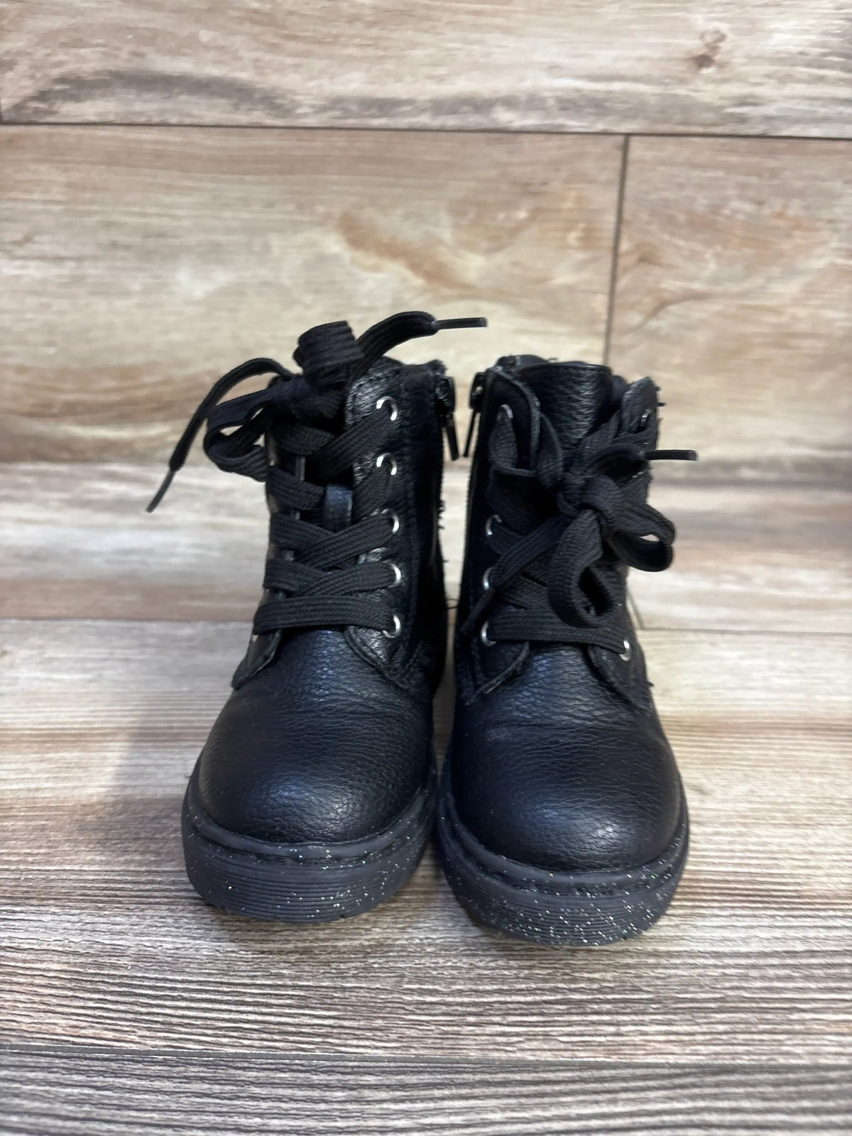 Front view of Sincerely Jules toddler black glitter lace-up boots showing glitter-speckled soles, side zippers, and textured leather on wood floor background
