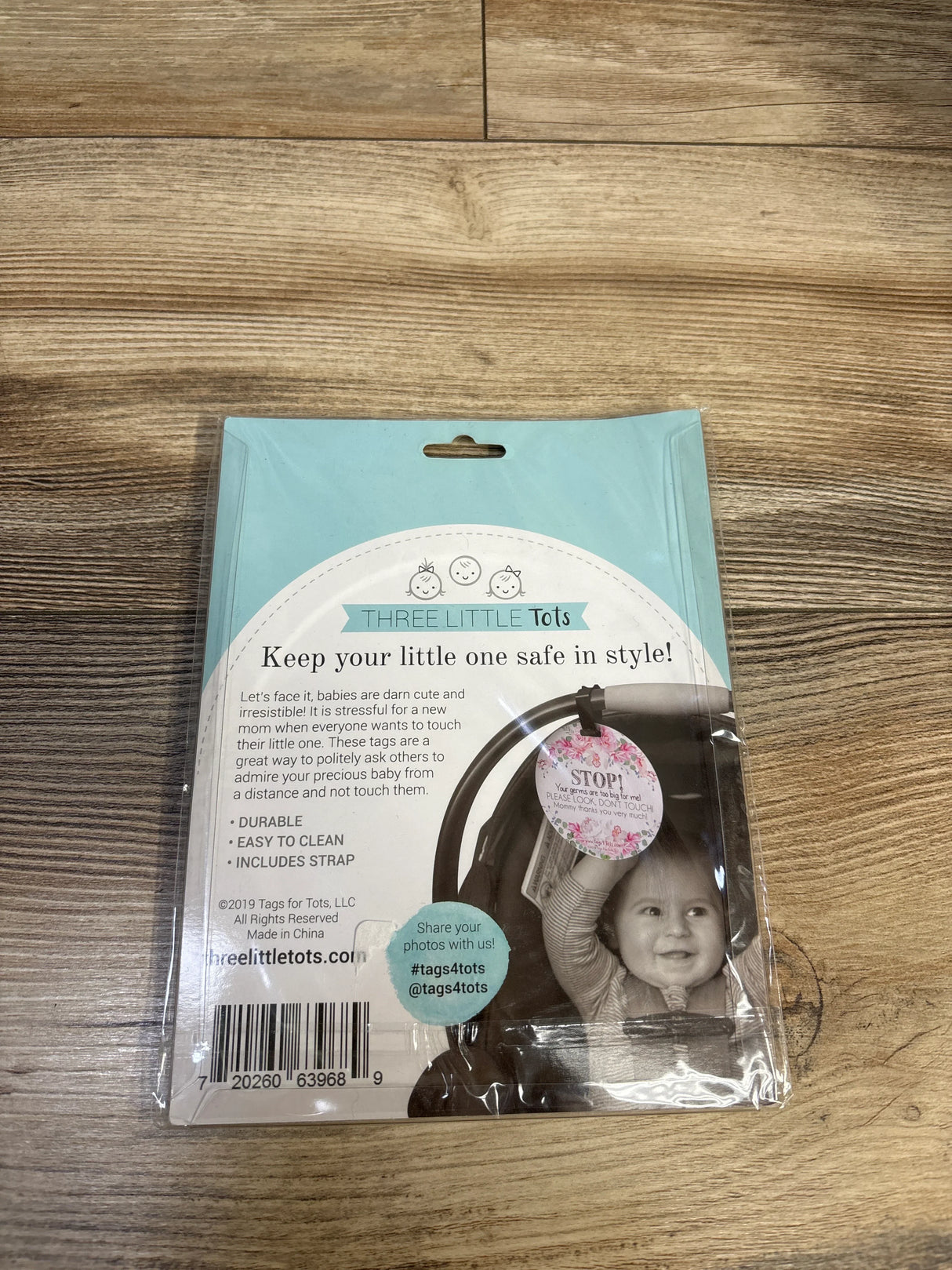 Front view of Three Little Tots car seat and stroller tag packaging showing floral ‘STOP’ tag and strap with ‘Keep your little one safe in style’ headline on wood surface