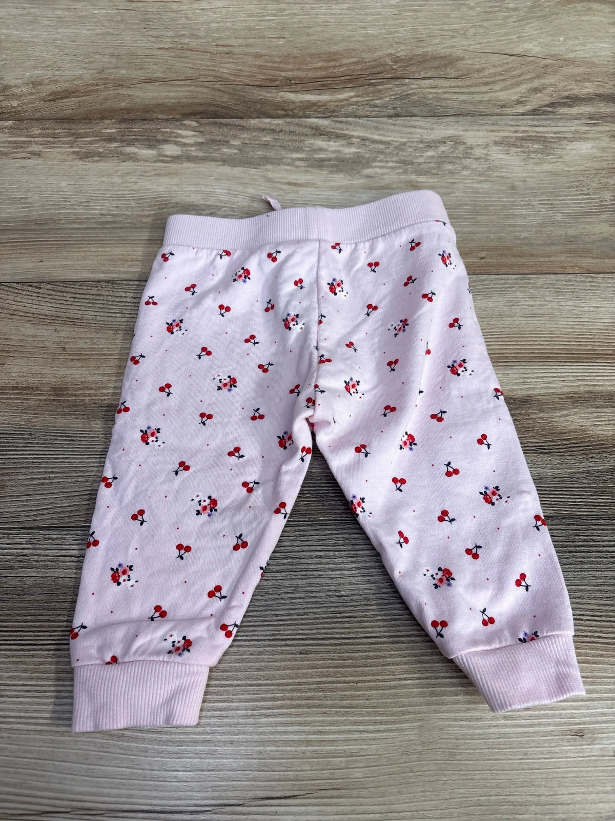 Flat lay of baby pink Janie & Jack French terry jogger pants showing red cherry print and ribbed cuffs on wood floor