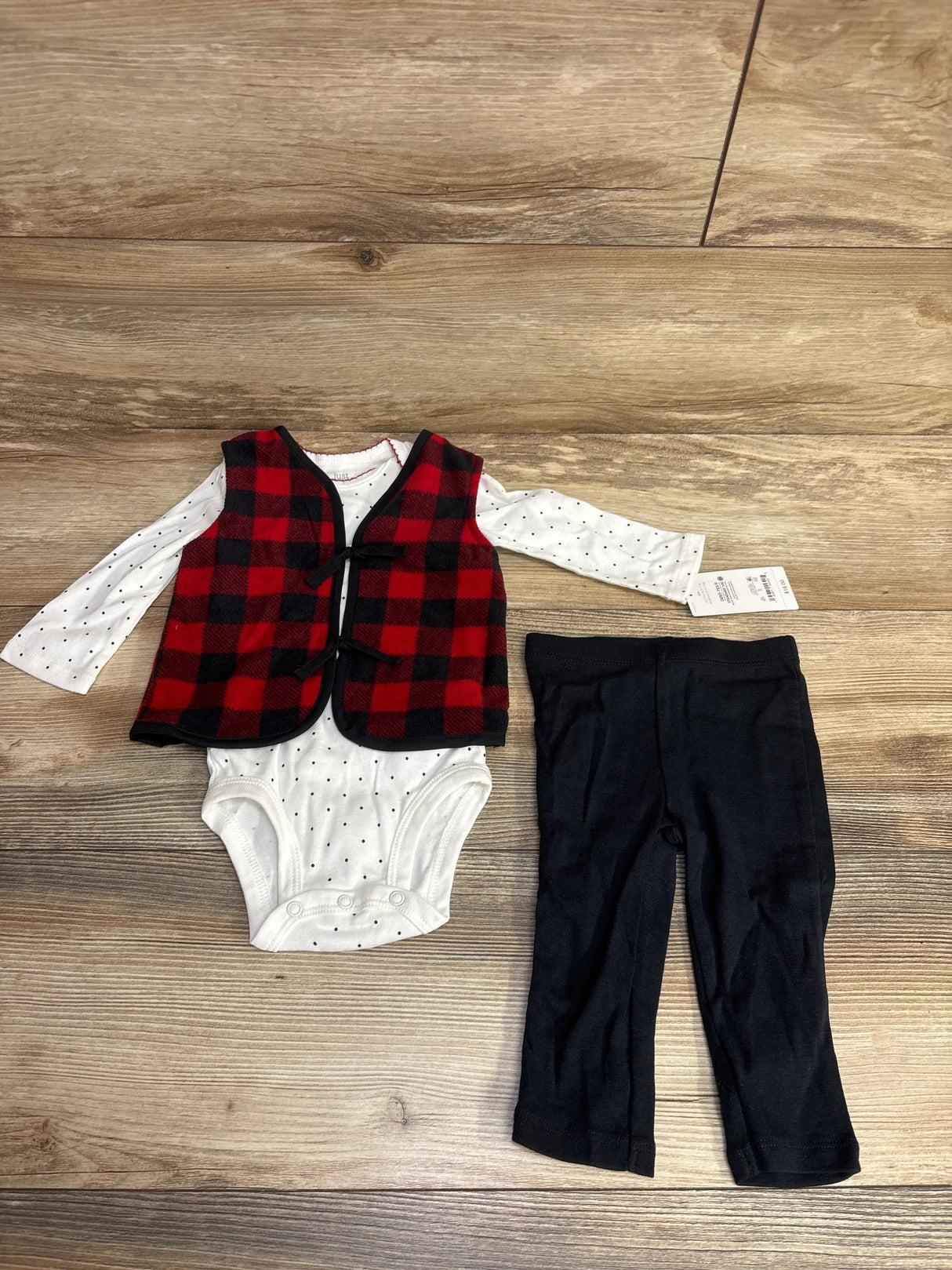 Baby outfit with red plaid vest, polka dot onesie, and black pants on wood floor