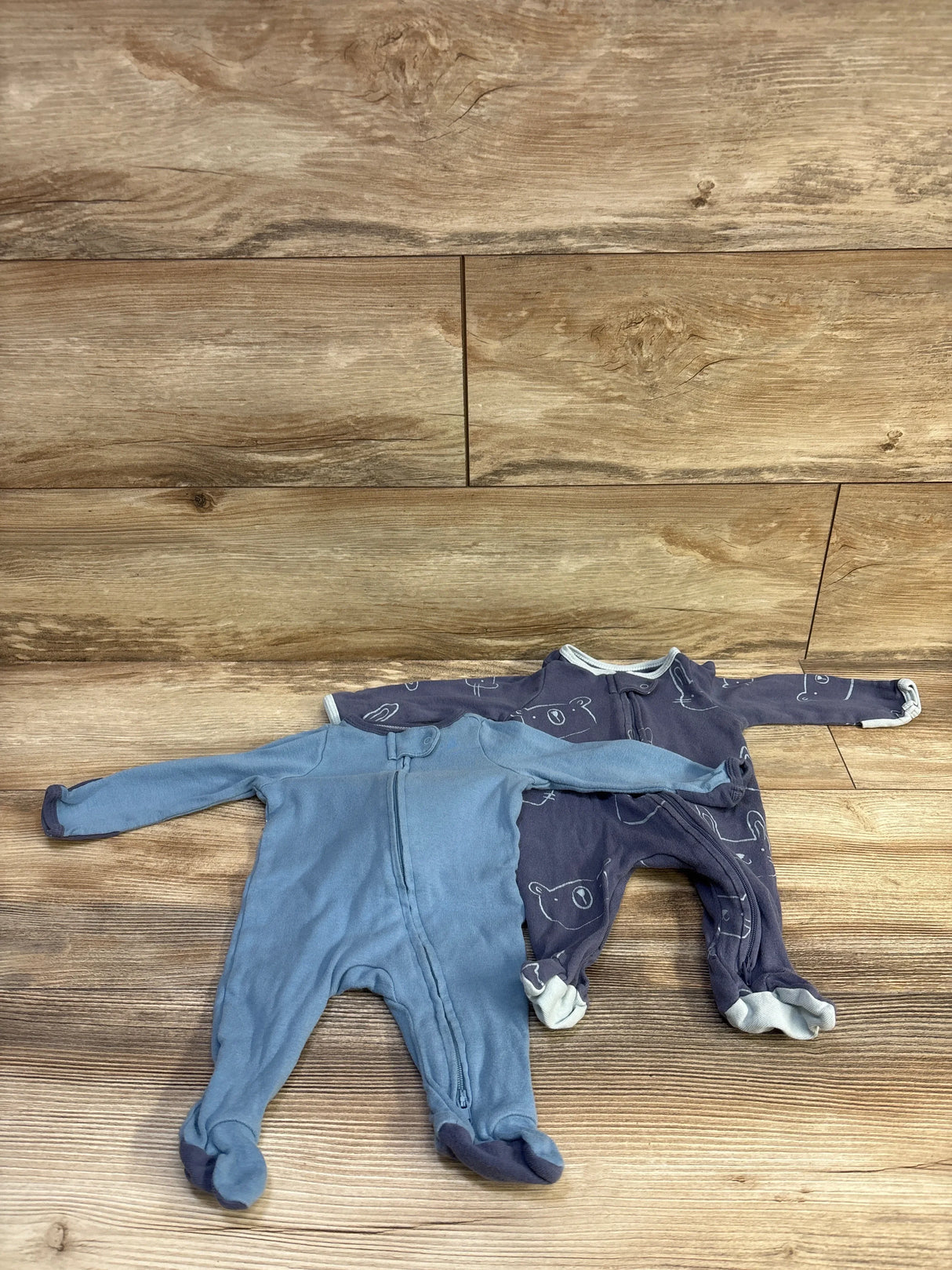 Flat lay of Cloud Island 2-pack newborn sleeper set (0-3M) showing front zipper, footed legs, blue solid and navy bear-print sleepers on wood floor background