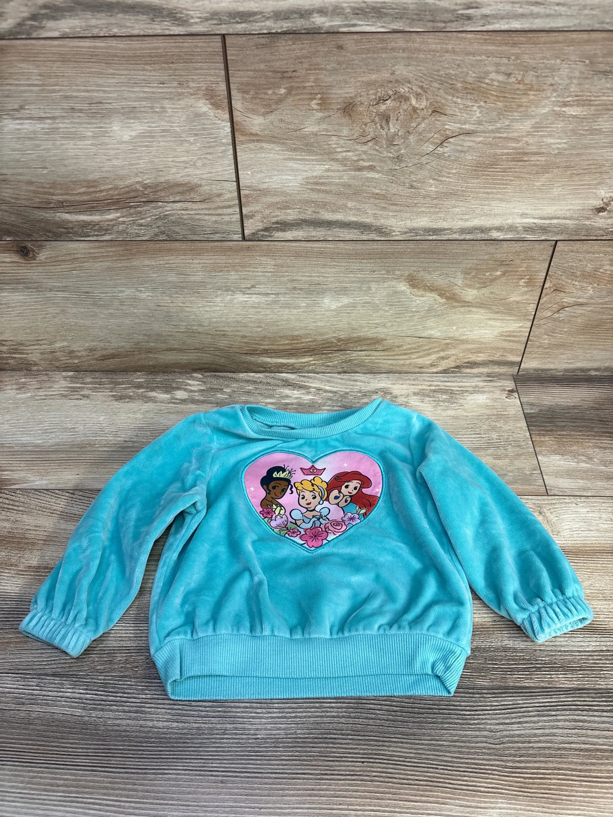 Front view of a teal Disney Princess sweatshirt showcasing a heart character graphic with a wood floor background