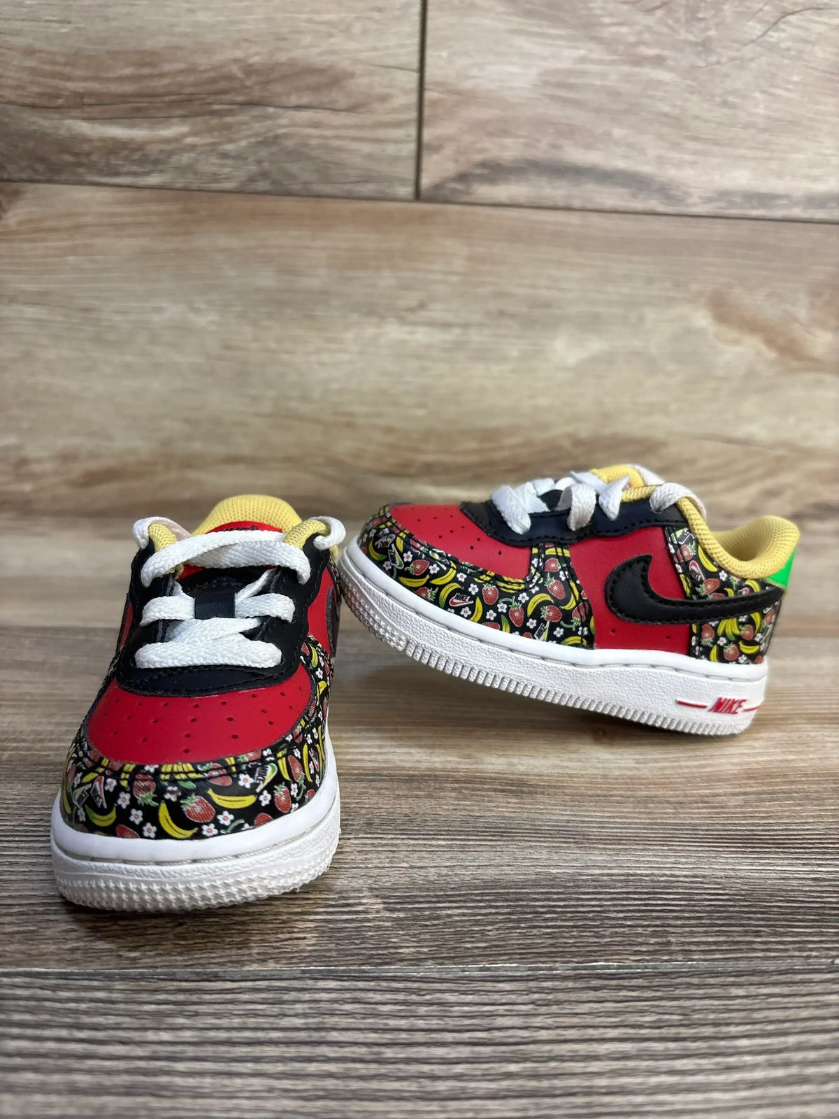 Front and side of Nike Force 1 TD Strawberry Banana shoes showing fruit-print panels, red toe and black Swoosh on wood floor