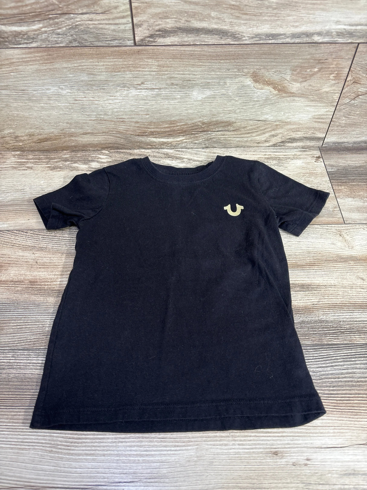 Black short sleeve kids t-shirt with a small gold logo on chest, laid on wood floor