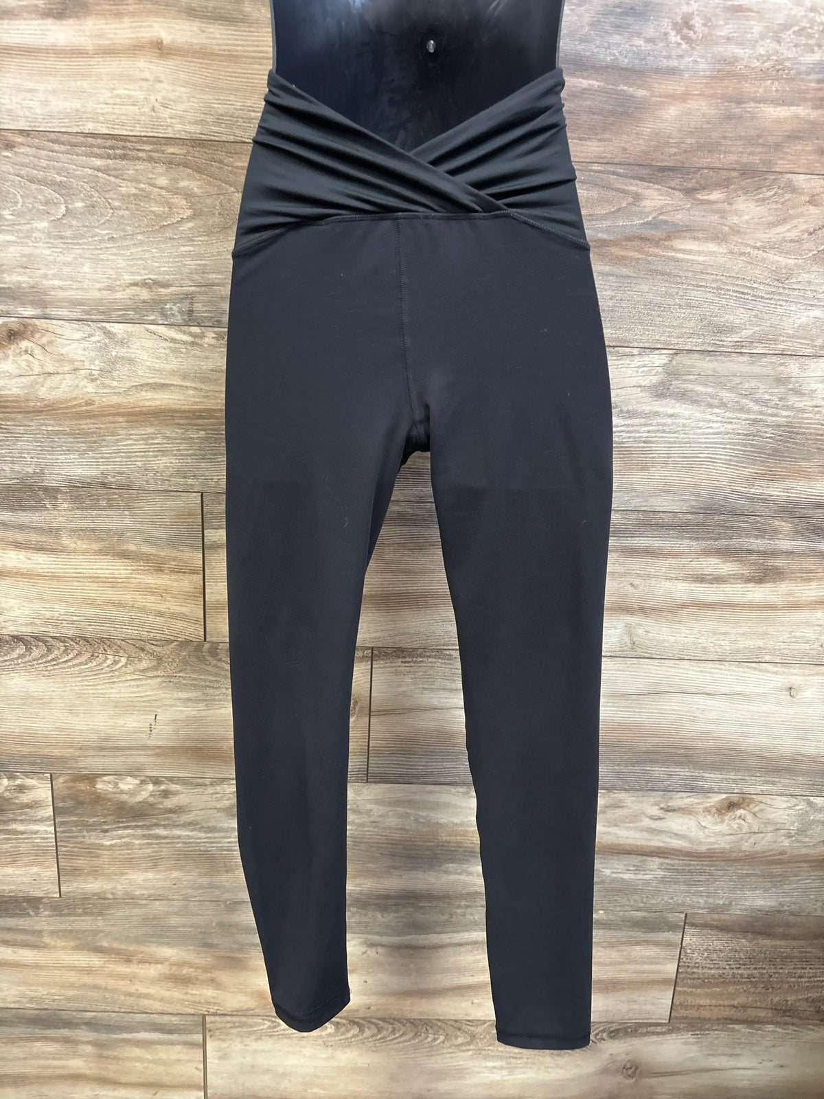 Front view of black maternity crossover panel leggings showcasing the crossed waistband with a wood plank background
