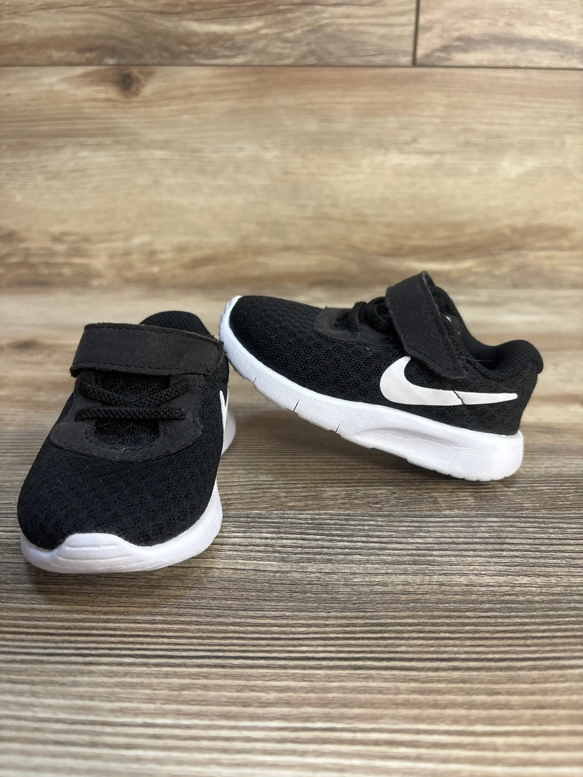 Front and side view of Nike Tanjun TDV toddler sneakers in black showing white soles, Velcro strap, and Nike swoosh on wood floor