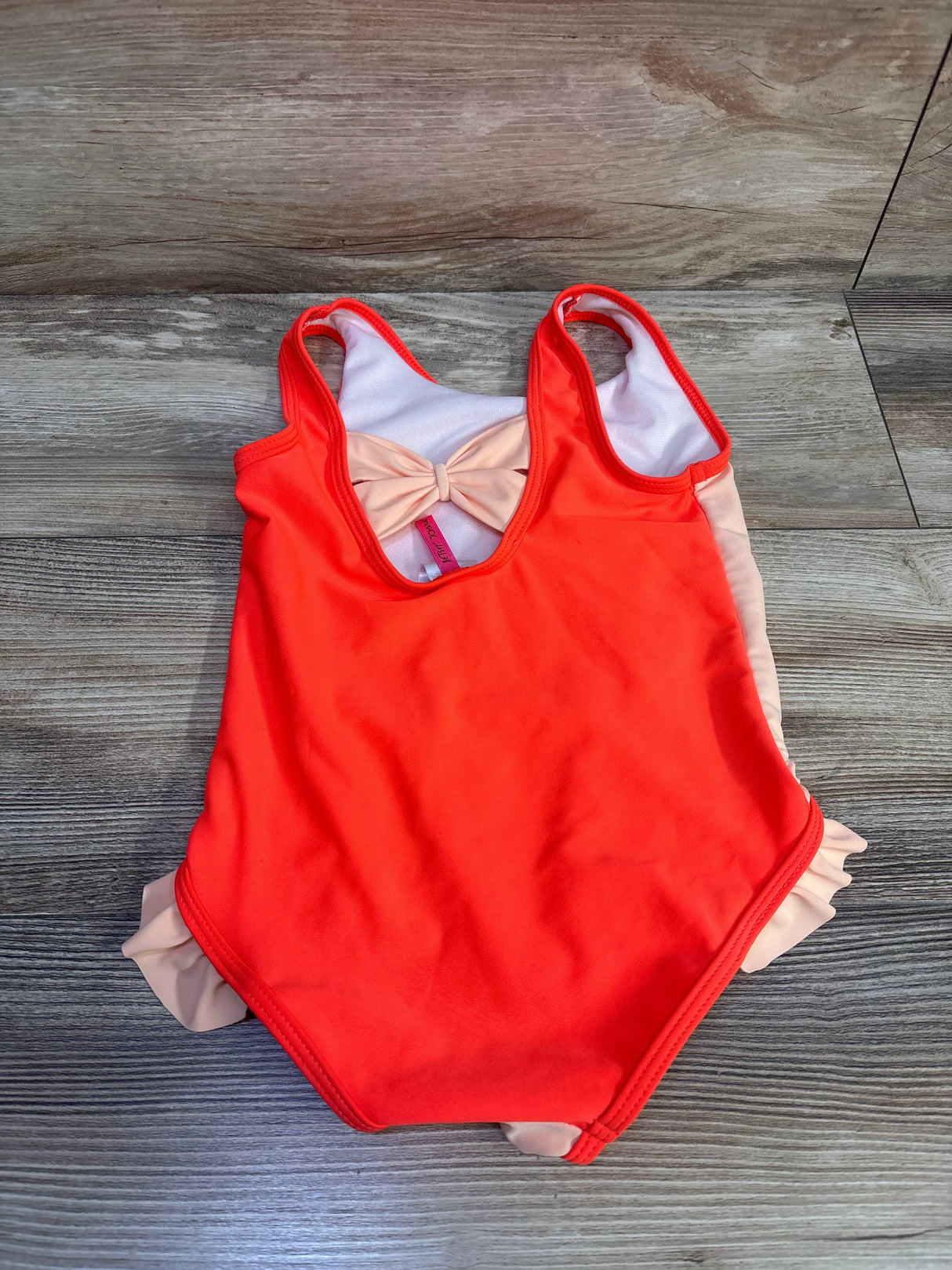 Flat lay of Betsey Johnson girls' one-piece swimsuit size 5T showcasing bright orange front, peach ruffle sides and back bow on wood floor