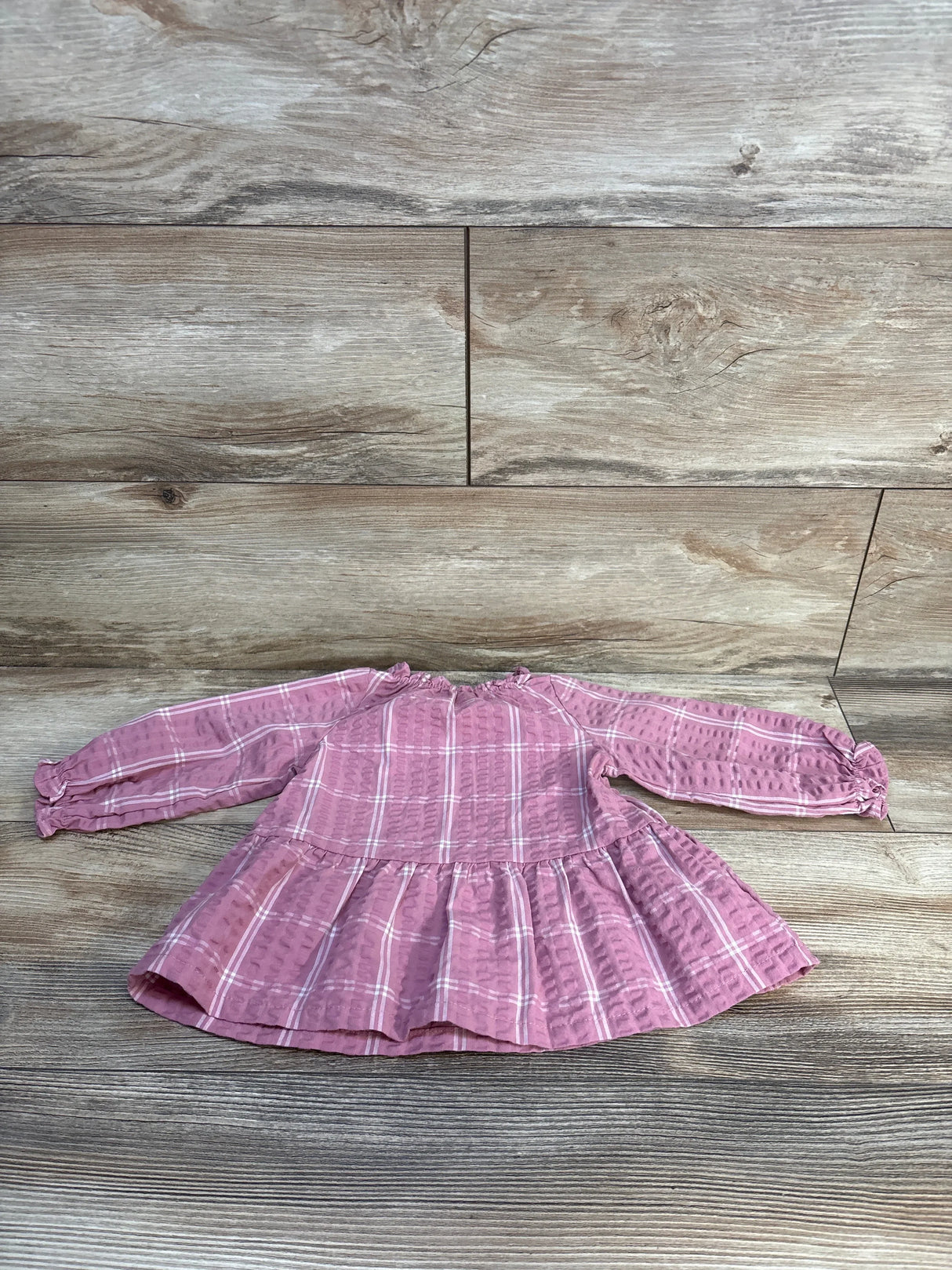 Pink plaid baby dress with long sleeves and ruffled hem, laid flat on wood floor