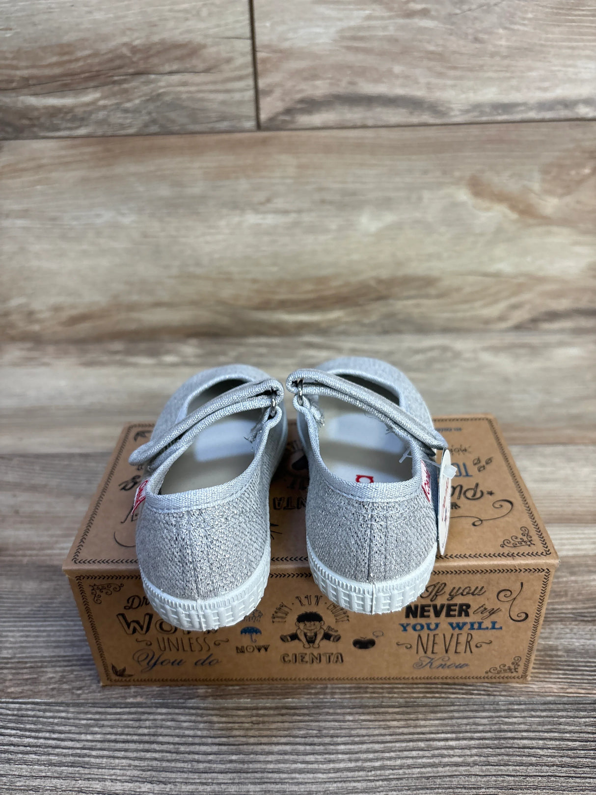 Rear view of Cienta Sparkle Mary Jane flats, silver toddler size 7C, showing glitter finish and Mary Jane strap on a decorative shoebox with wood background