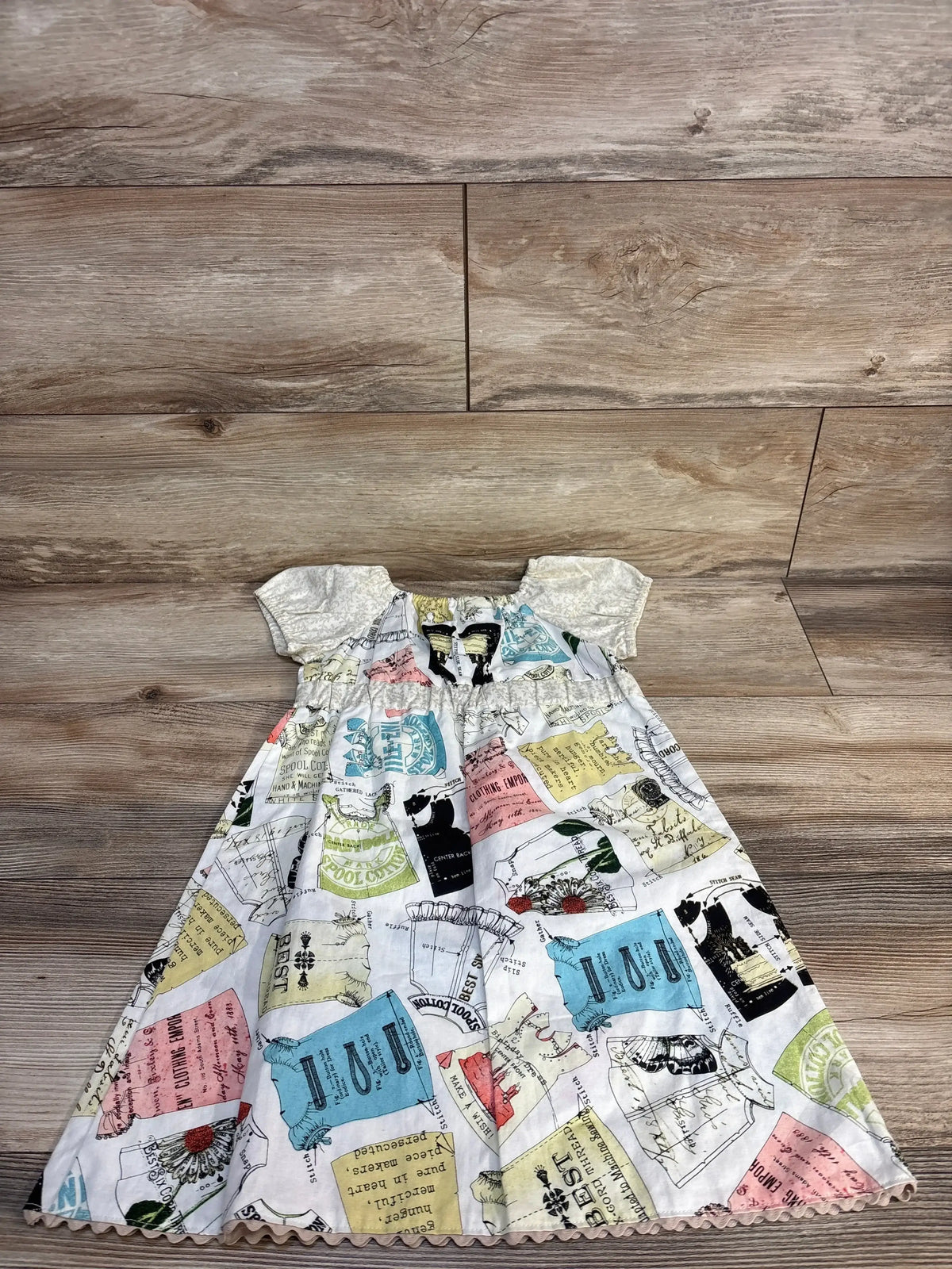 Vintage-style baby dress with sewing pattern print, ruffled sleeves, and scalloped hem on wood floor
