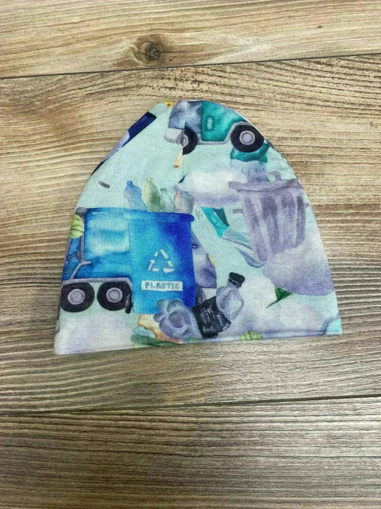 Flat lay of Posh P baby beanie size 0-3 months showing teal and blue recycling truck and plastic bin print on wood background