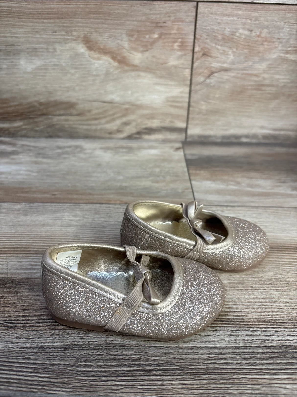 Angled side view of Koala Kids gold glitter bow ballet flats showing glitter finish, bow and elastic strap on wood background