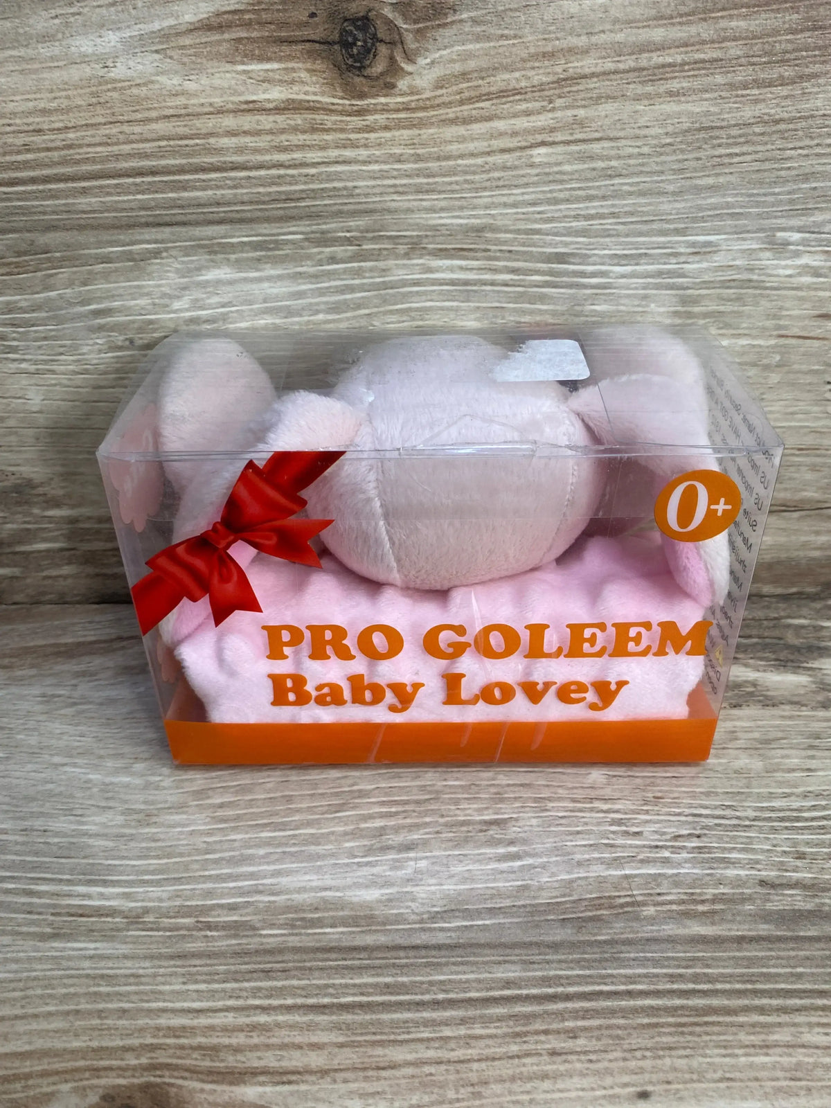 Front view of NEW Pro Goleem Baby Lovey pink bunny security blanket in original clear gift box packaging with red bow on wood plank floor