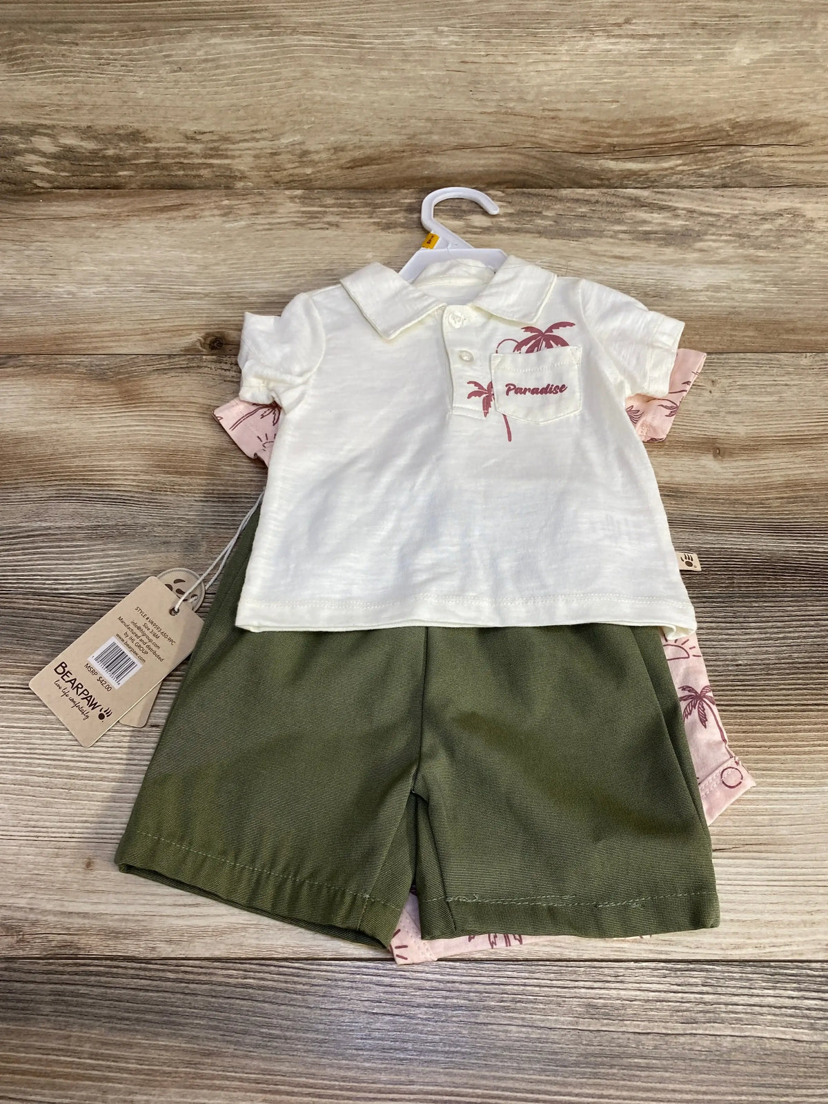 Flat lay of Bear Paw 3-piece baby short set showing white polo with "Paradise" chest print, pink palm-print undershirt, and olive shorts on wooden background