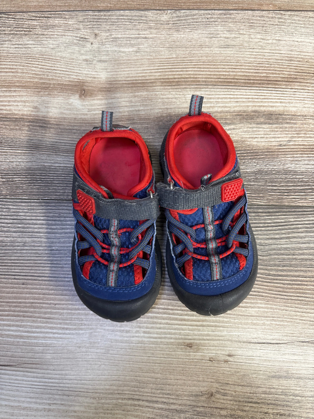Top-down of Oshkosh Pumba2 closed-toe toddler sandals, navy and red, showing bungee laces, gray Velcro strap and black rubber toe caps on wood floor