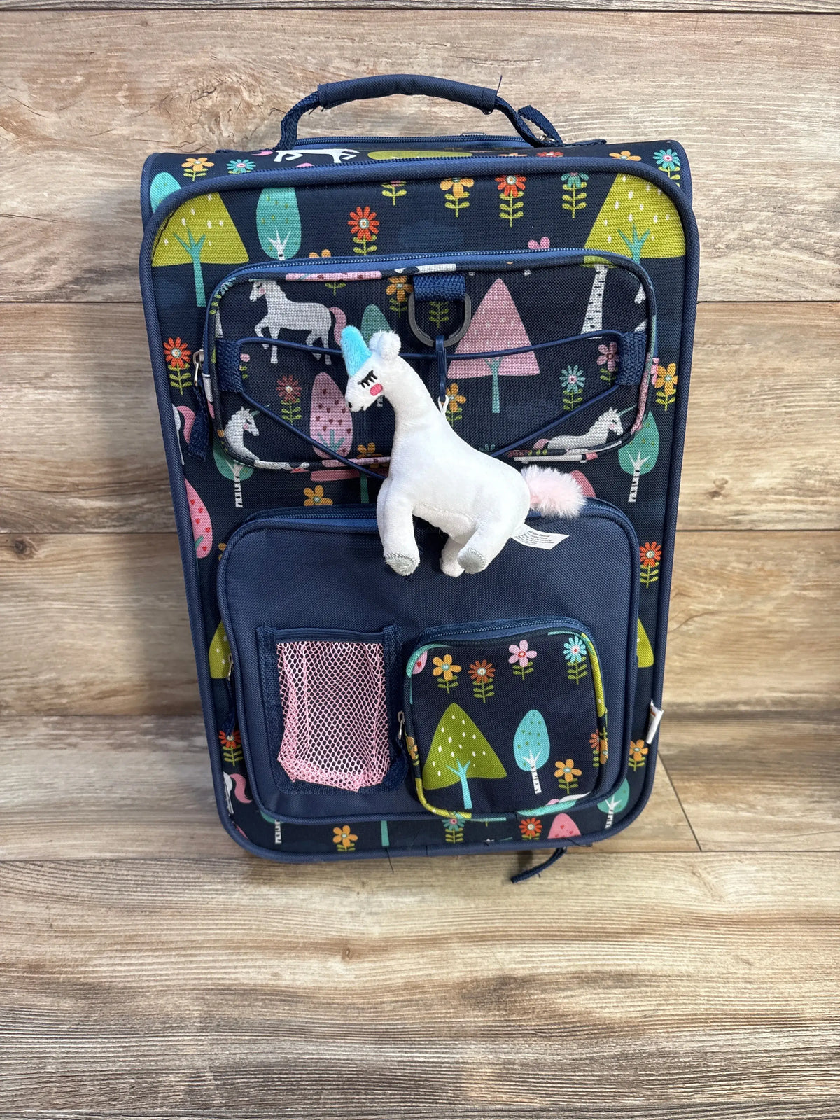 Front view of Crckt Kids’ softside carry-on suitcase in unicorn design with zippered pockets and bottle holder on wood floor