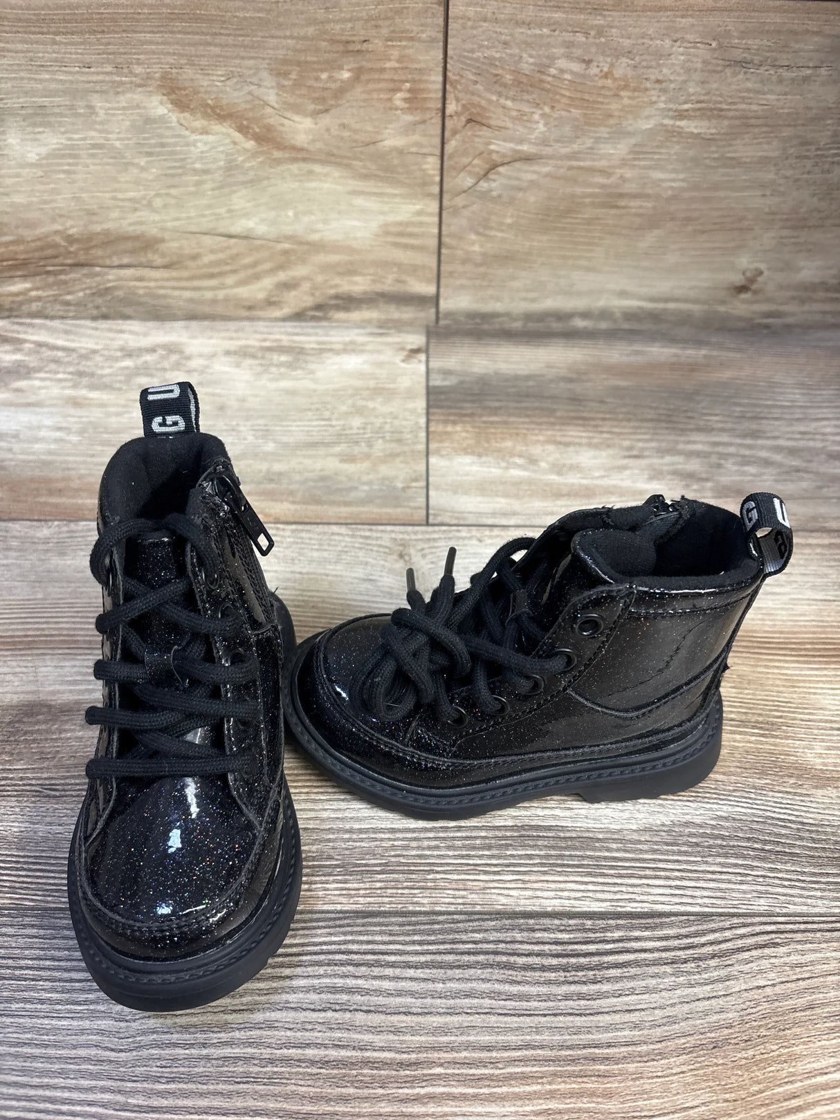 Front-left view of UGG Kids Robley black glitter toddler boots showing glitter finish, laces and side zipper on wood floor