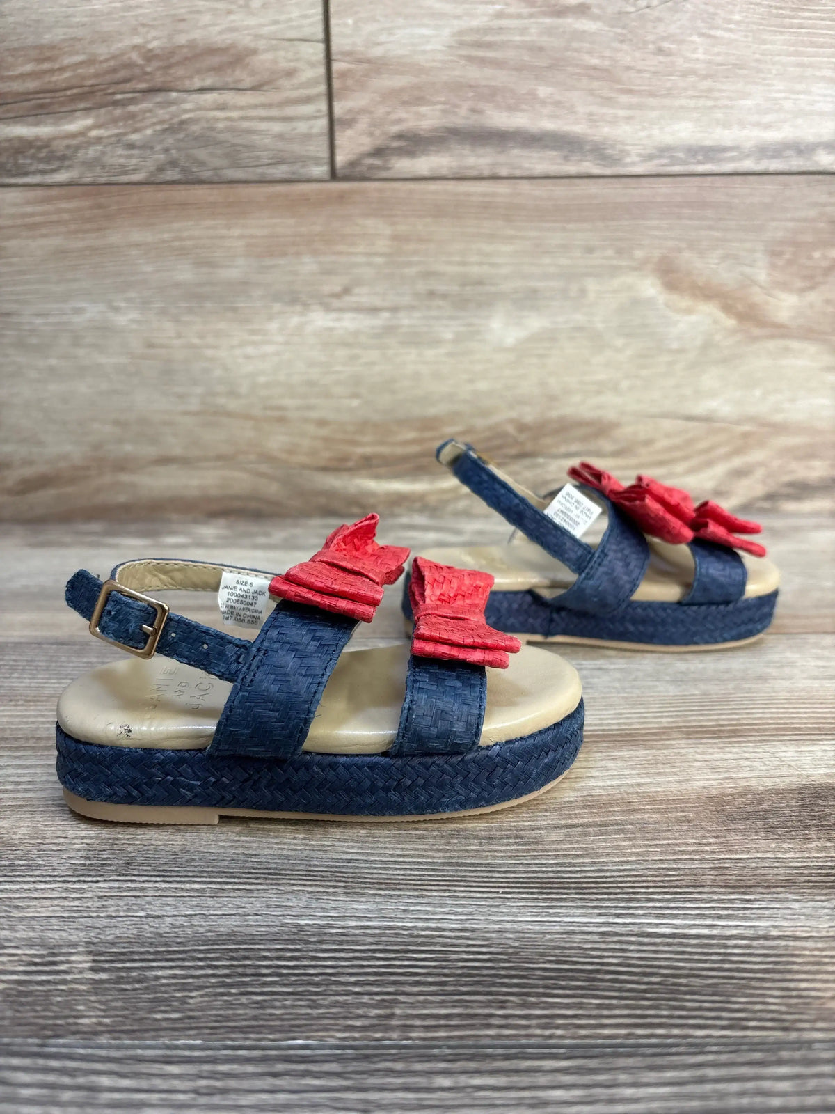 Three quarter view of Janie and Jack double bow straw sandals in navy blue with red bows and woven sole on wood plank floor
