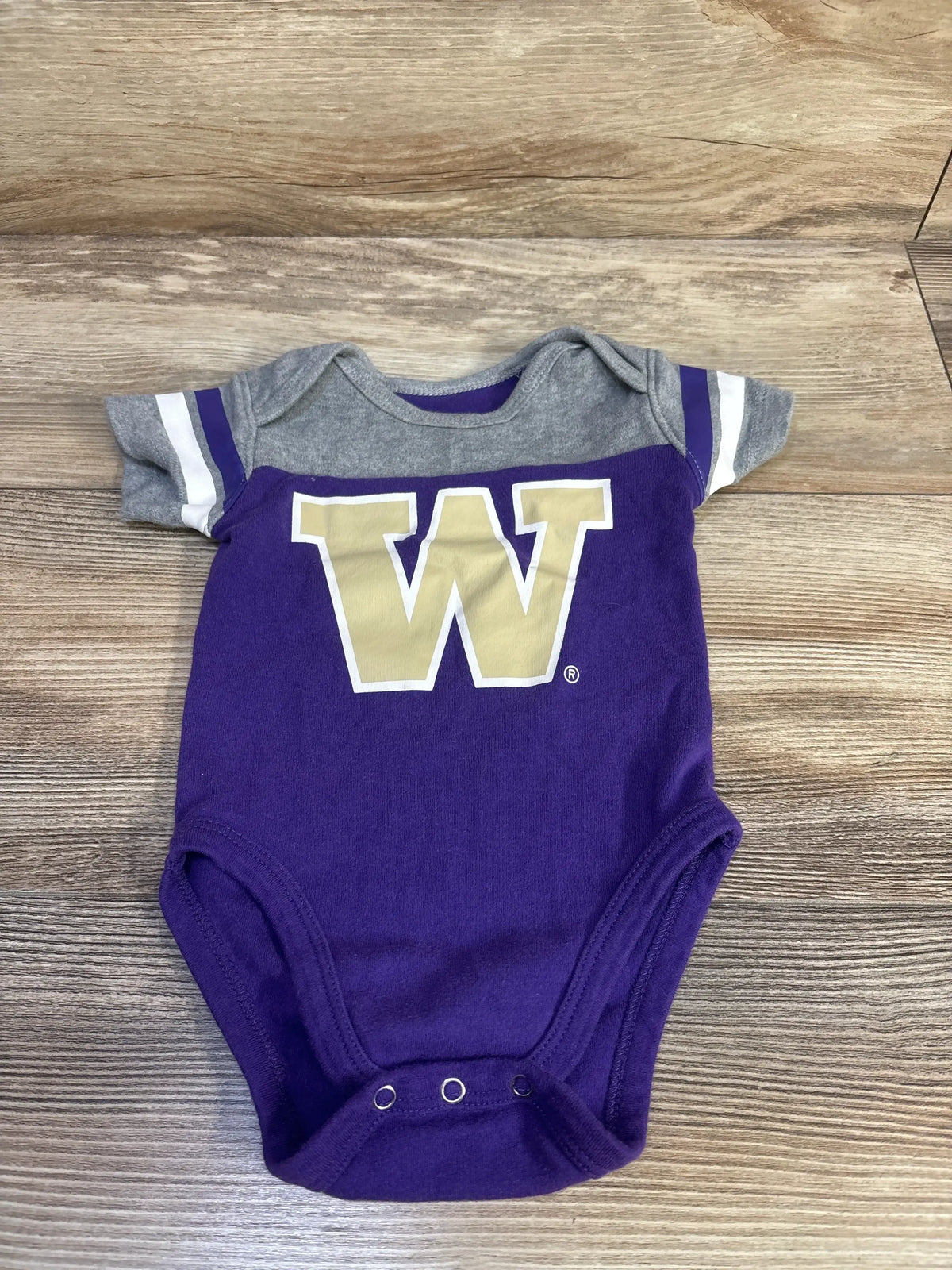 Front view of UW baby bodysuit with gold W, purple body, gray striped sleeves and three snaps on wood background