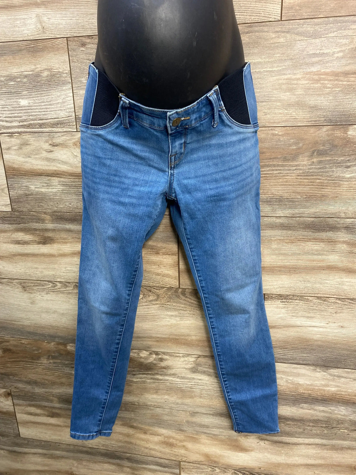 Front view of blue maternity jeggings showcasing elastic side panels, button-and-zip fly, and tapered ankle-length legs on wood-plank background