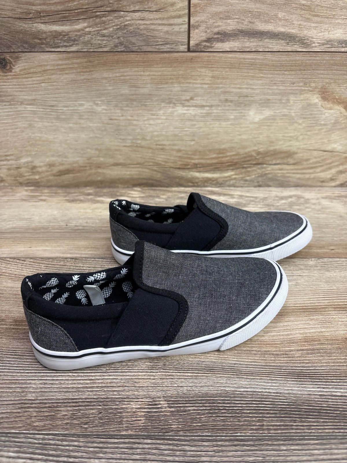 Side view of Cat & Jack gray canvas slip-on sneakers showcasing black elastic panels and pineapple-print lining on wood flooring
