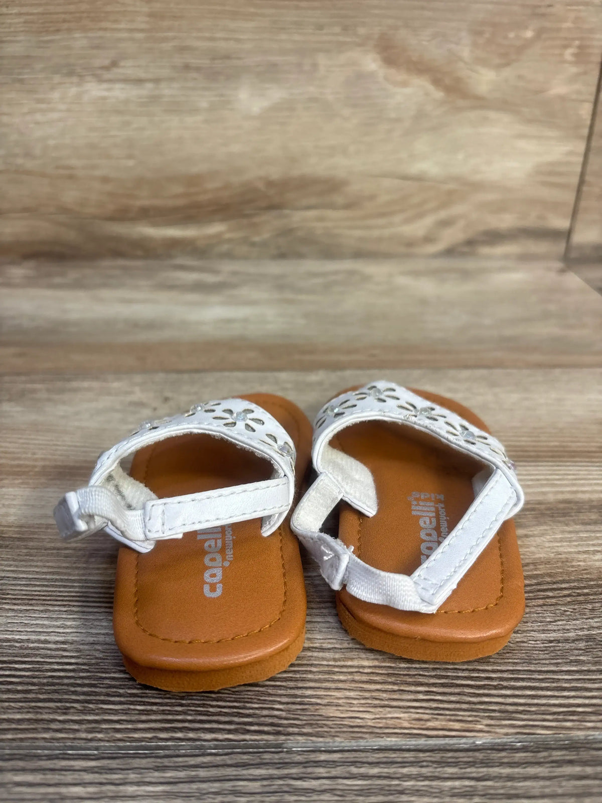 Top view of Capelli white backstrap sandals showcasing perforated uppers and brown footbeds on a wood grain floor