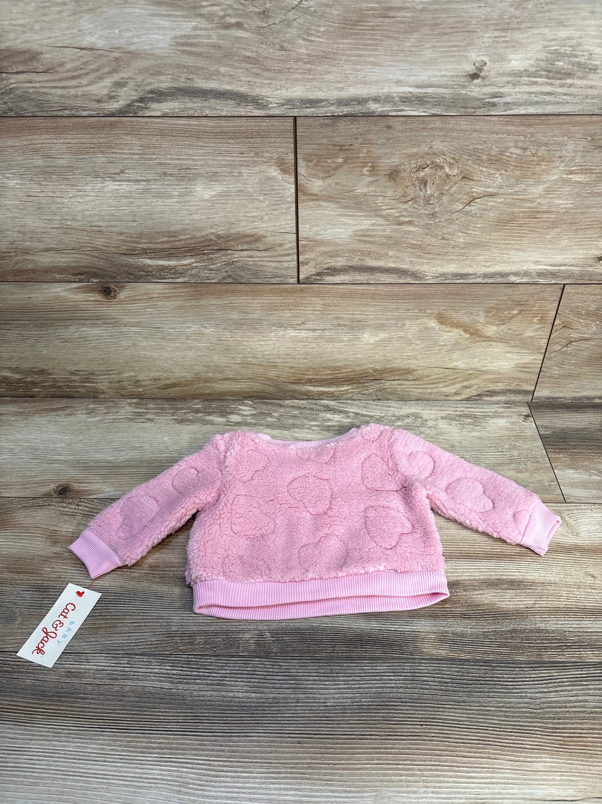 Pink textured baby sweater with embossed hearts, long sleeves, and ribbed cuffs on wood background