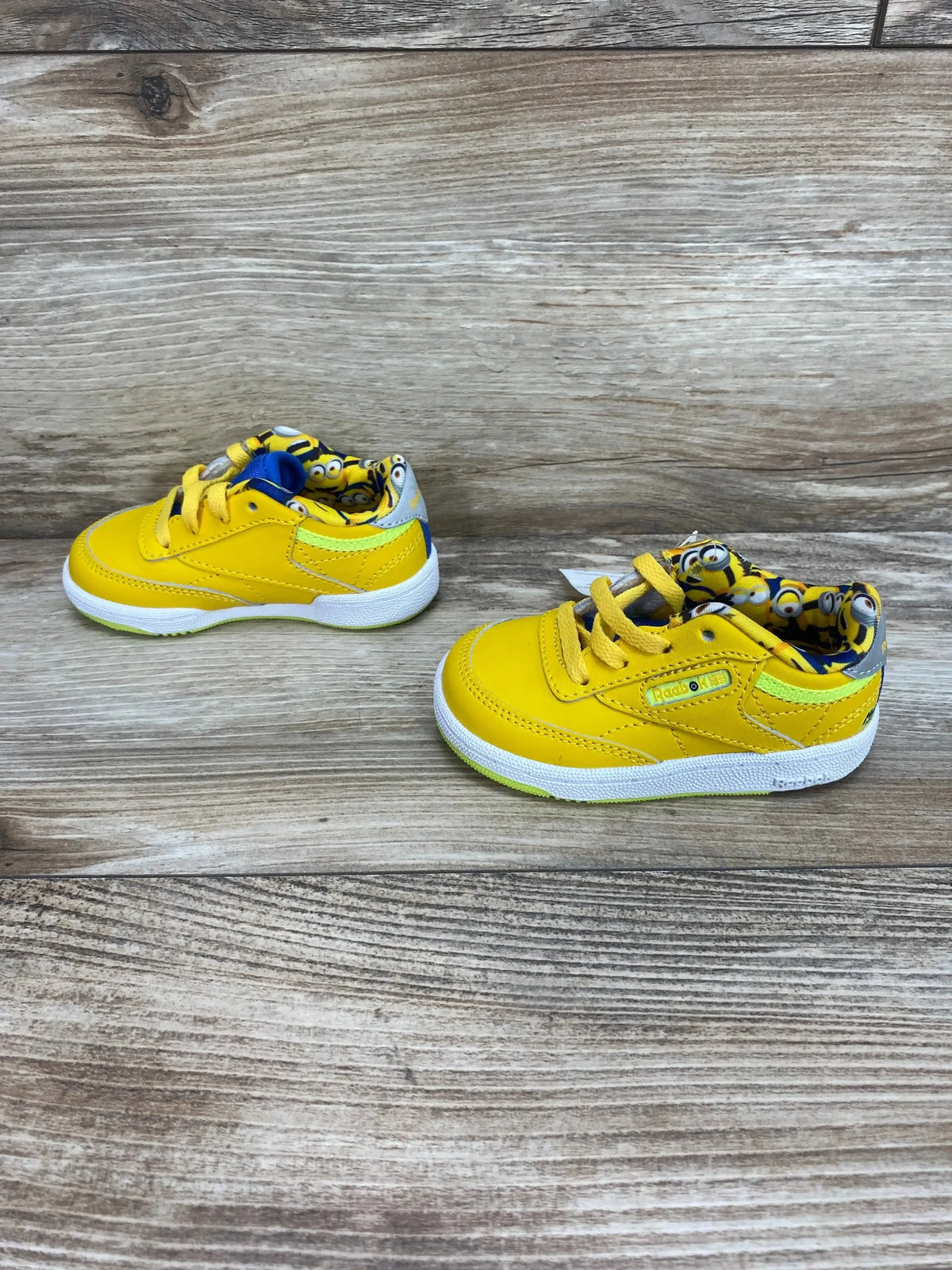 Angled side view of toddler yellow Minions Club C 85 sneakers showing Minions-print inner lining and white sole on wood background