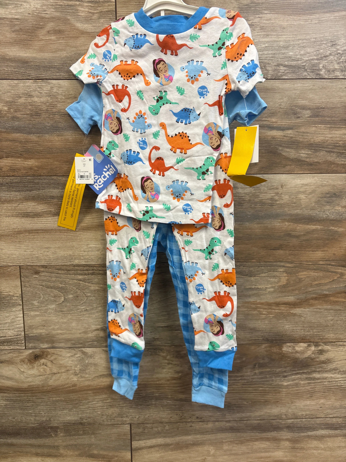 Front view of Ms Rachel 4-piece toddler pajama set showing colorful dinosaur and Ms Rachel portrait print, blue cuffed sleeves and blue checked leggings on wood floor
